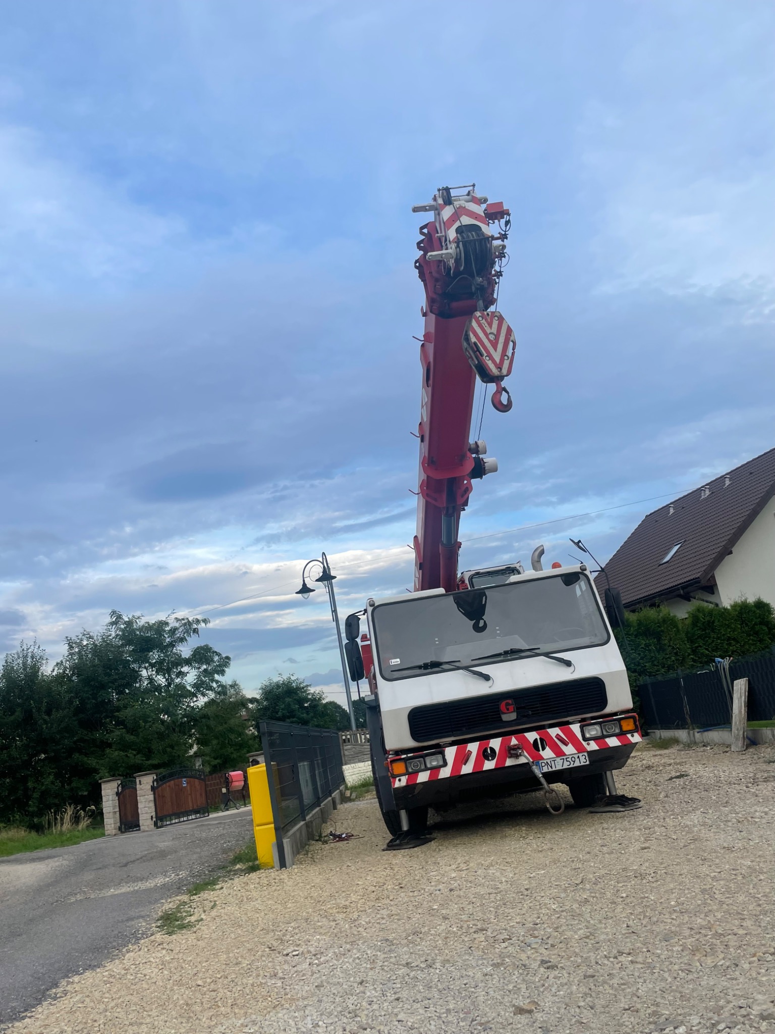Ciężki, biało-czerwony dźwig samochodowy z wysuniętym ramieniem na tle pochmurnego nieba i zabudowań.
