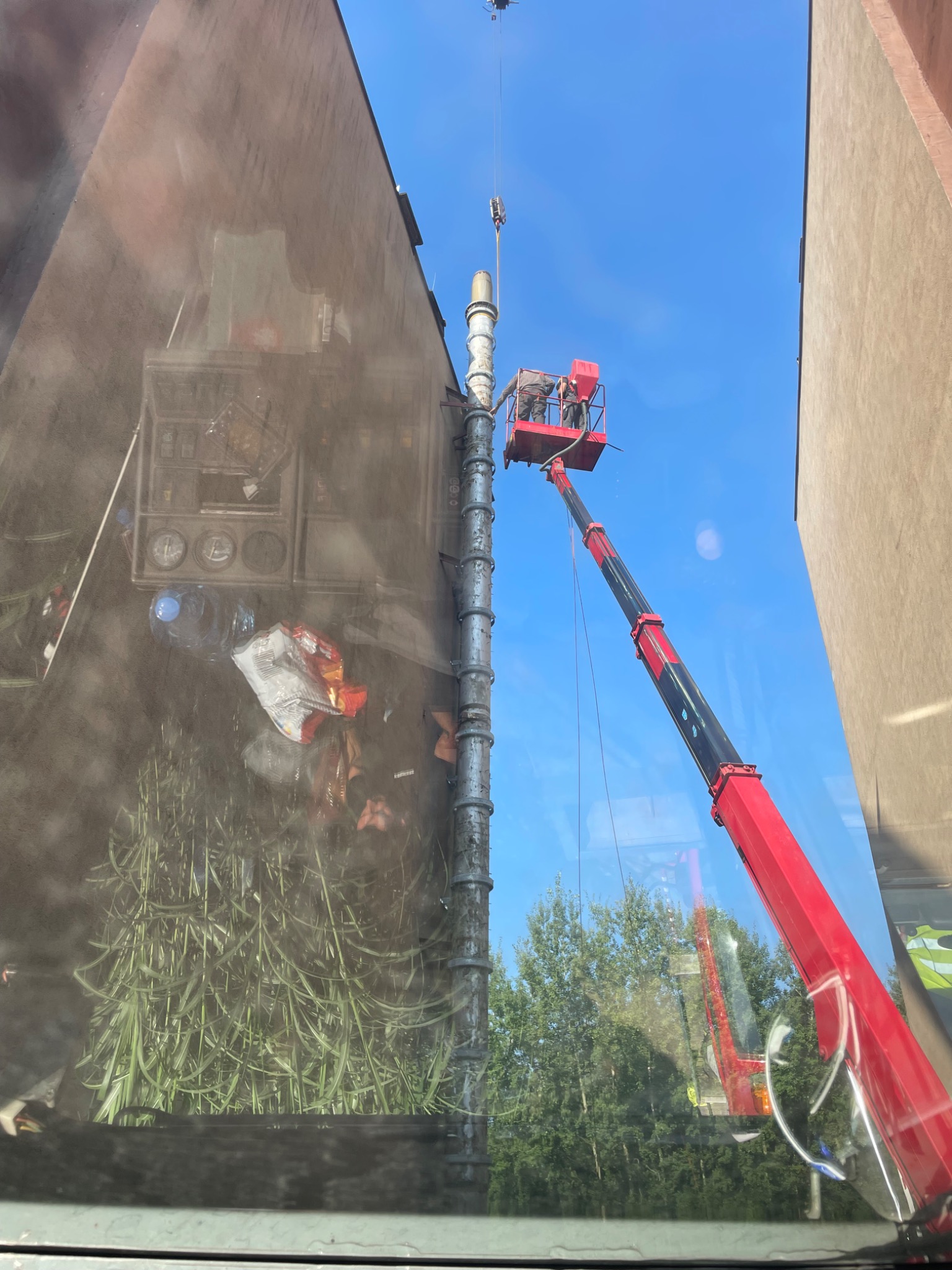 Prace wysokościowe z użyciem podnośnika koszowego przy montażu komina, widok zza szyby z odbiciem wnętrza pojazdu.