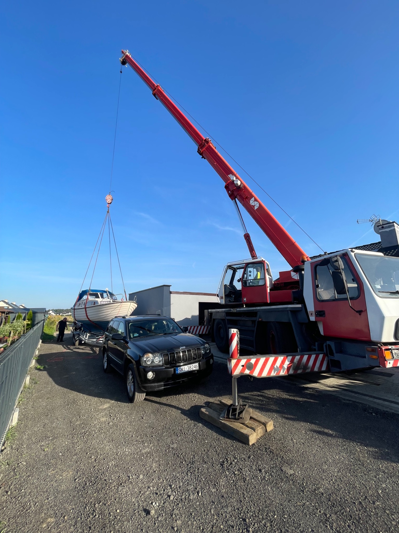Czerwono-biały dźwig samochodowy podnosi łódź na przyczepie, obok czarny Jeep Grand Cherokee z widoczną rejestracją, błękitne niebo w tle.