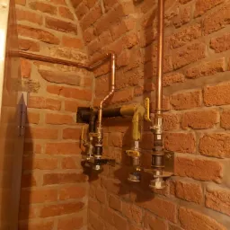 Miedziane rury instalacji wodnej z zaworami zamontowane na ścianie z cegły, widoczne fragmenty armatury i przyłącza.
