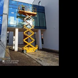 Pracownik w żółtej kamizelce i kasku, na podnośniku koszowym, myje szklaną fasadę nowoczesnego budynku biurowego z licznymi odbiciami nieba.