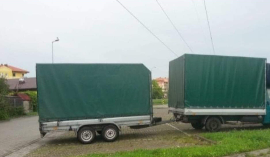 Ciężarówka z przyczepą z zielonymi plandekami na parkingu, widoczne budynki w tle.