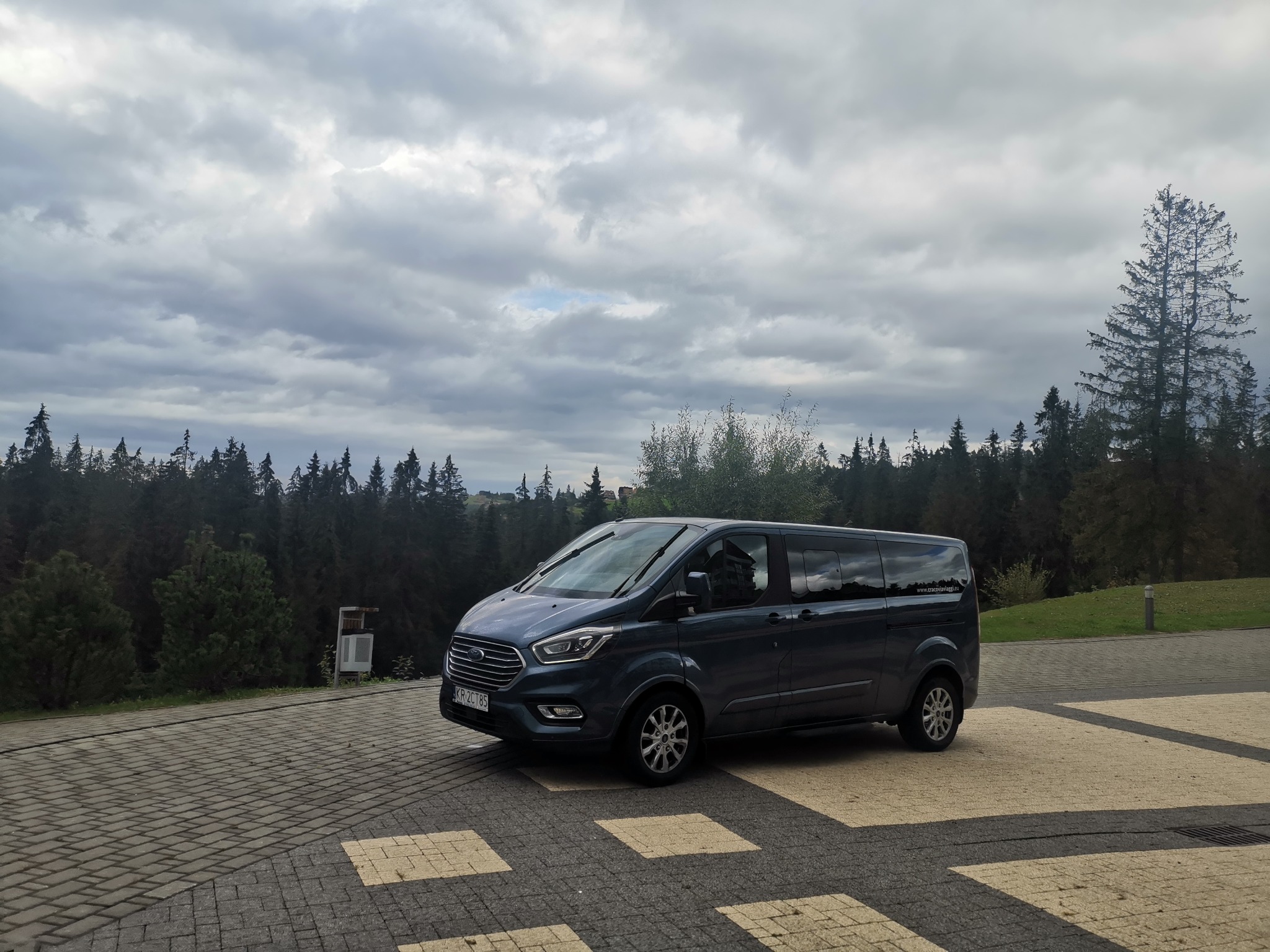 Granatowy bus Ford Transit Custom na parkingu z kostki brukowej, w tle las iglasty i pochmurne niebo.