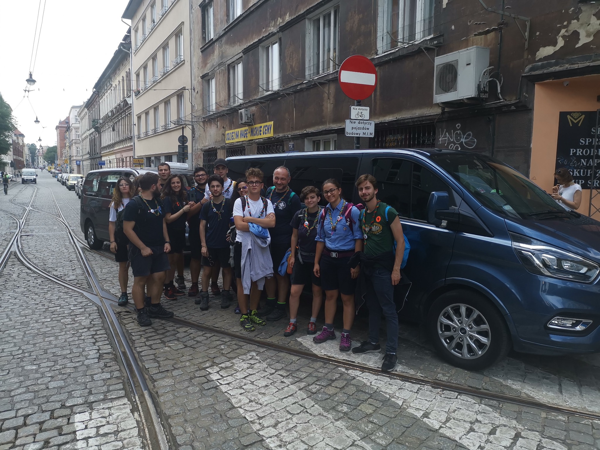 Grupa harcerzy w pełnym umundurowaniu pozuje przed ciemnoniebieskim busem na tle krakowskiej ulicy z torami tramwajowymi.