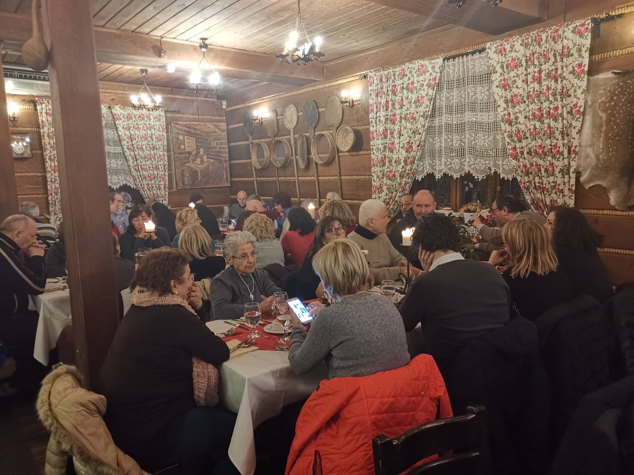 Grupa osób siedzi przy stołach w góralskiej restauracji z drewnianymi ścianami ozdobionymi tradycyjnymi przedmiotami i wzorzystymi zasłonami.