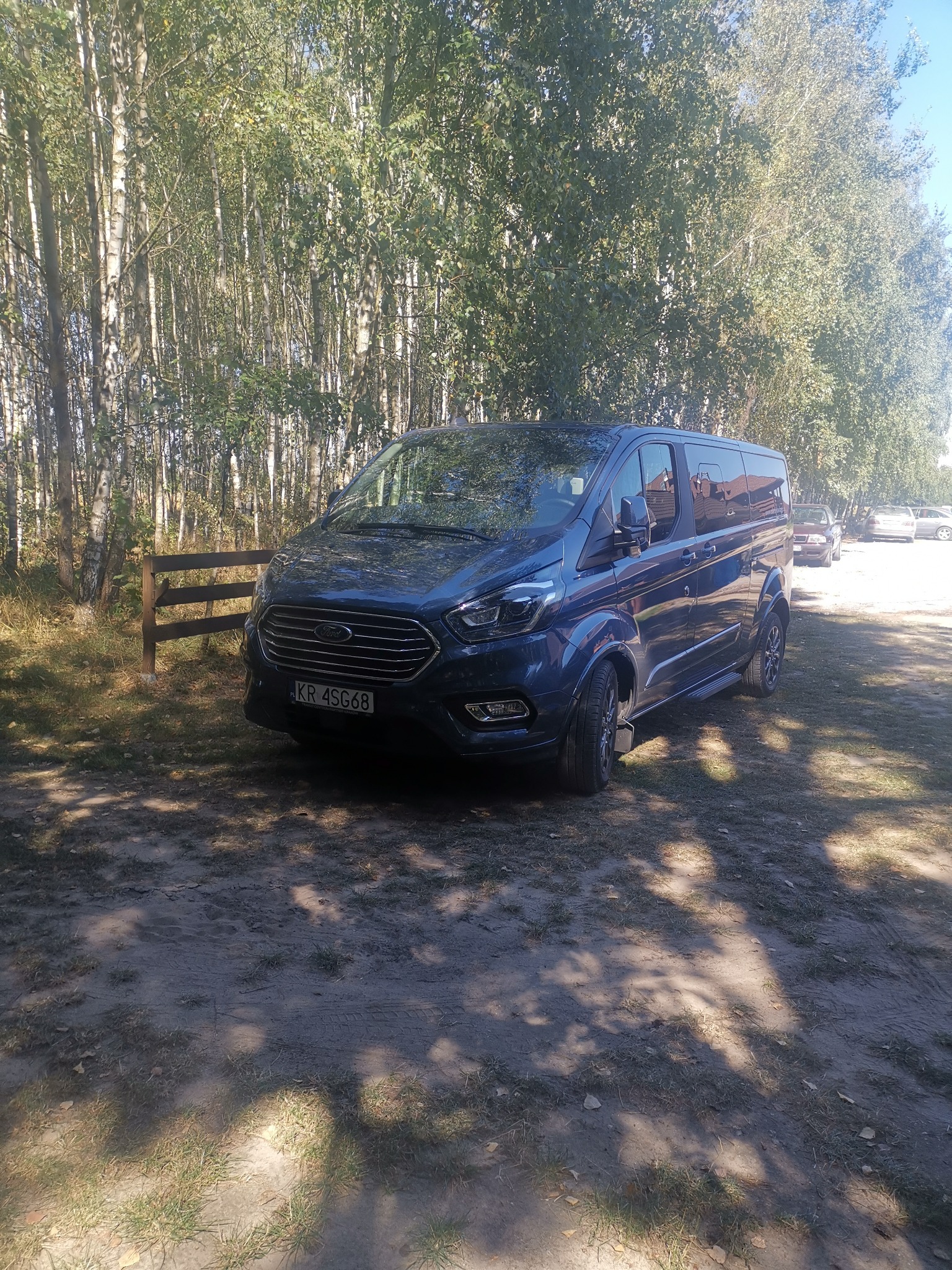 Niebieski Ford Transit Custom parkuje na piaszczystym terenie w cieniu drzew, widoczny fragment drewnianego płotu i inne pojazdy w tle.