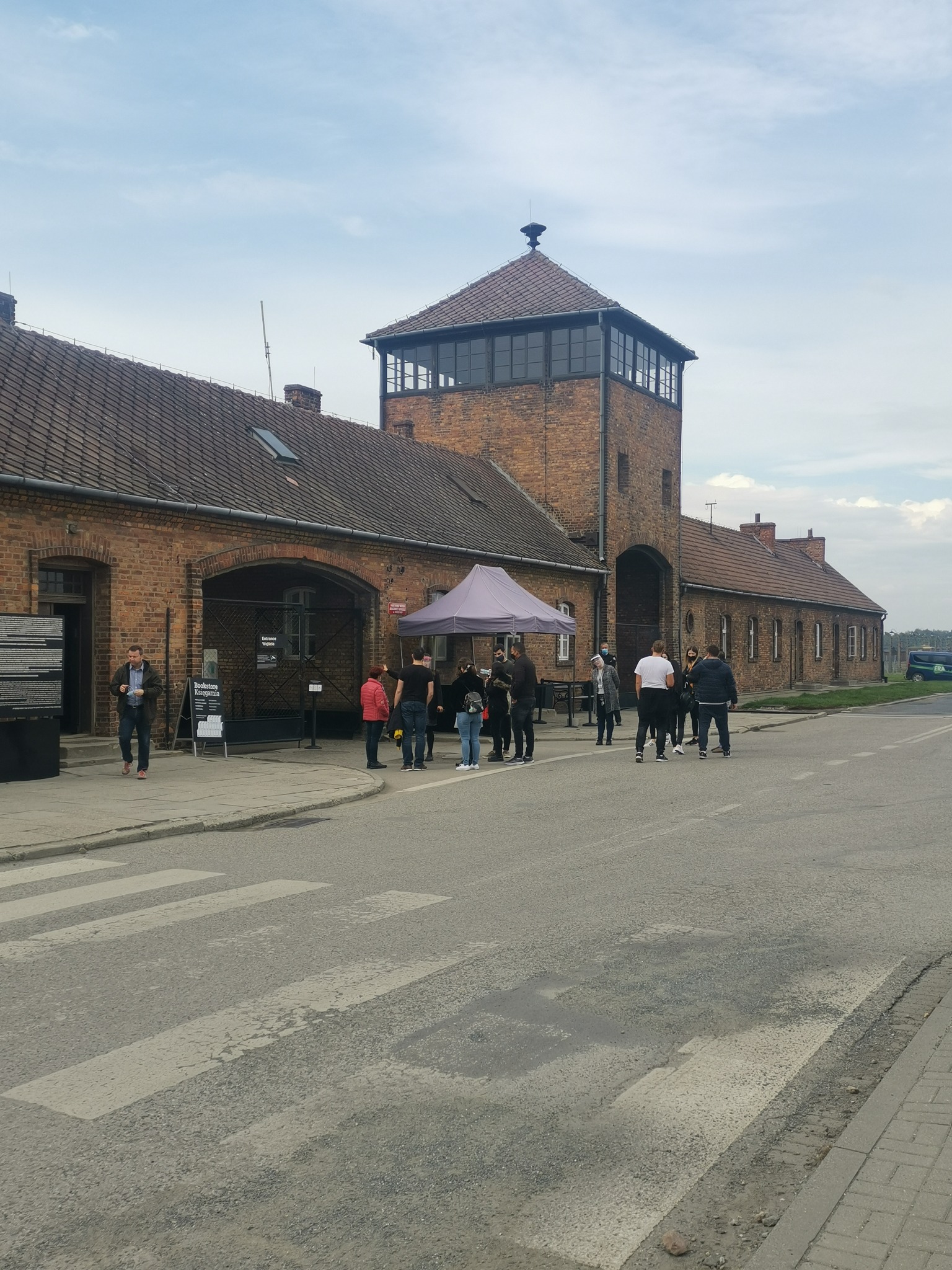 Brama wjazdowa do obozu Auschwitz-Birkenau z grupą turystów oczekujących na wejście, widoczny fragment tablicy informacyjnej i zaparkowany bus z boku.