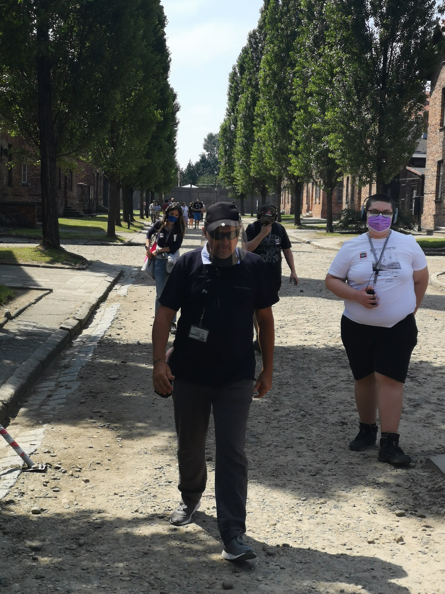 Grupa turystów idzie brukowaną drogą między budynkami z czerwonej cegły, otoczona wysokimi drzewami, część osób nosi maseczki.