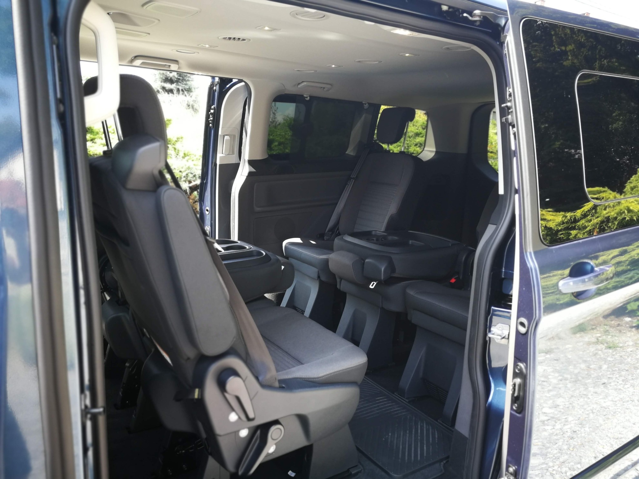 Wnętrze ciemnoniebieskiego busa z rozsuwanymi drzwiami, widoczne szare fotele z podłokietnikami i zagłówkami, oraz czarna gumowa wycieraczka na podłodze.