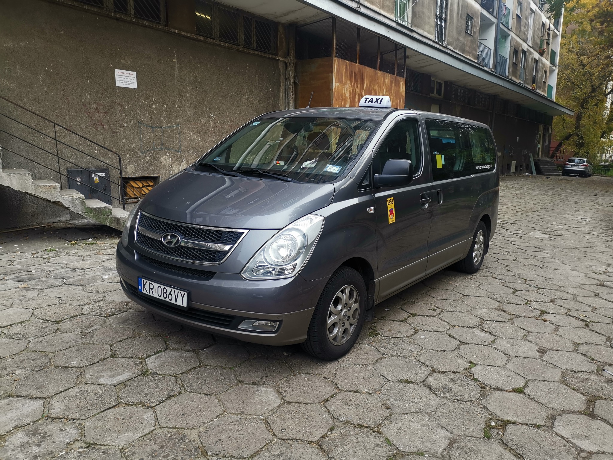 Szary minivan taxi Hyundai z tablicą rejestracyjną KR 086VY, stojący na betonowej kostce brukowej przed szarym budynkiem.