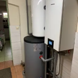 Zestaw grzewczy z dwoma zbiornikami i panelem sterowania Innova, zainstalowany w piwnicy z terakotą na podłodze i zielonymi drzwiami w tle.