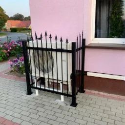 Biała jednostka zewnętrzna klimatyzacji, zabezpieczona czarnym, ozdobnym ogrodzeniem, umieszczona na kostce brukowej przy różowej ścianie budynku z oknem.