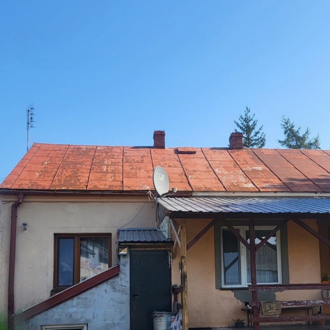 Dom jednorodzinny z czerwoną, częściowo odbarwioną dachówką, anteną satelitarną i widocznymi kominami na tle błękitnego nieba.