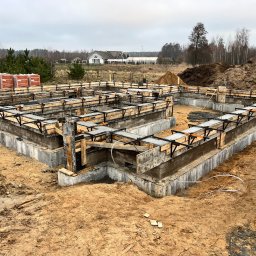 ZAW TECH - Wylany fundament z szalunkiem drewnianym i zbrojeniami, widoczne cegły na paletach, w tle dom jednorodzinny i drzewa.