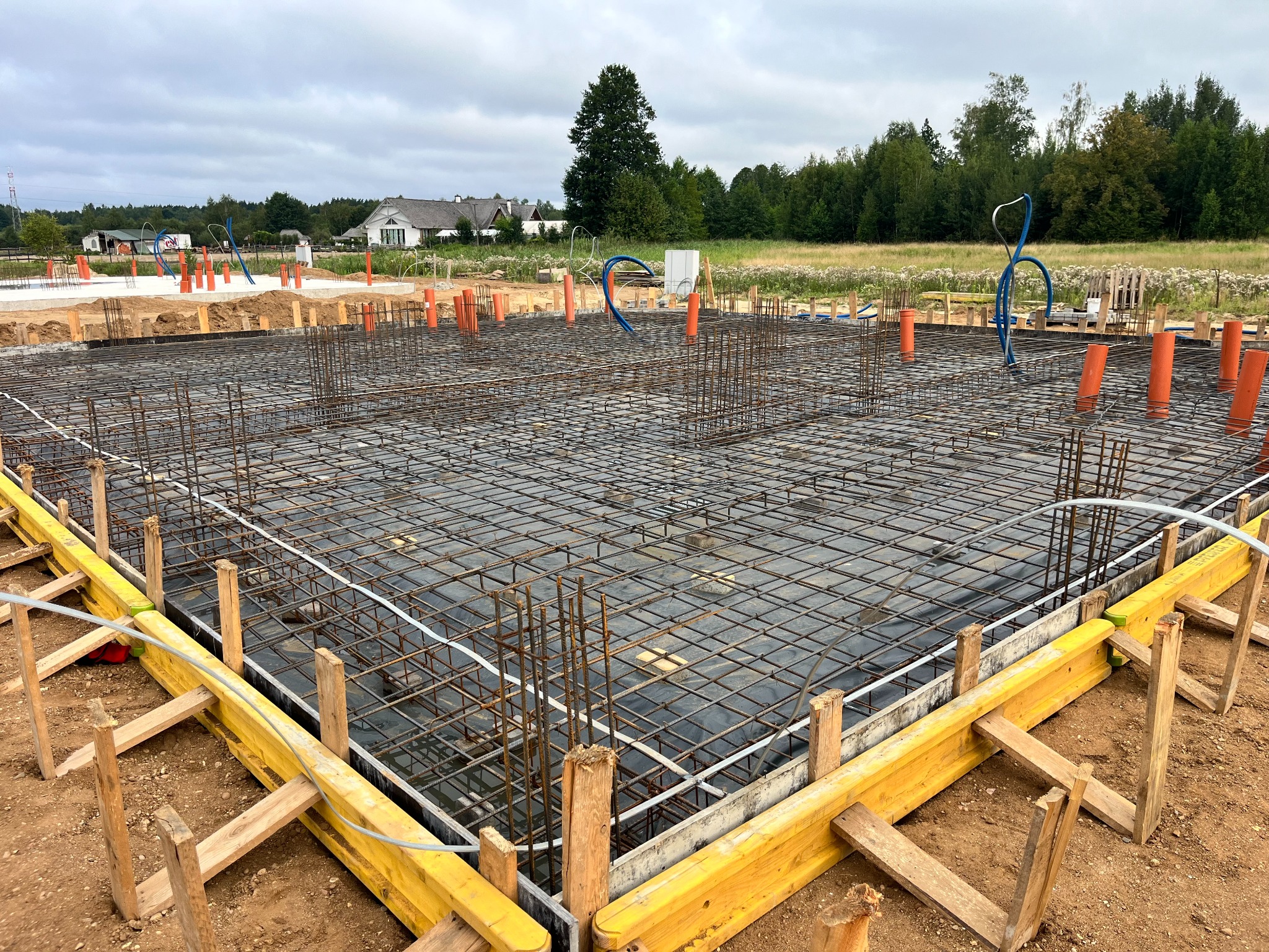 Zbrojenie fundamentów budynku jednorodzinnego z widocznymi rurami instalacyjnymi i żółtym szalunkiem, na tle zieleni i zabudowań.