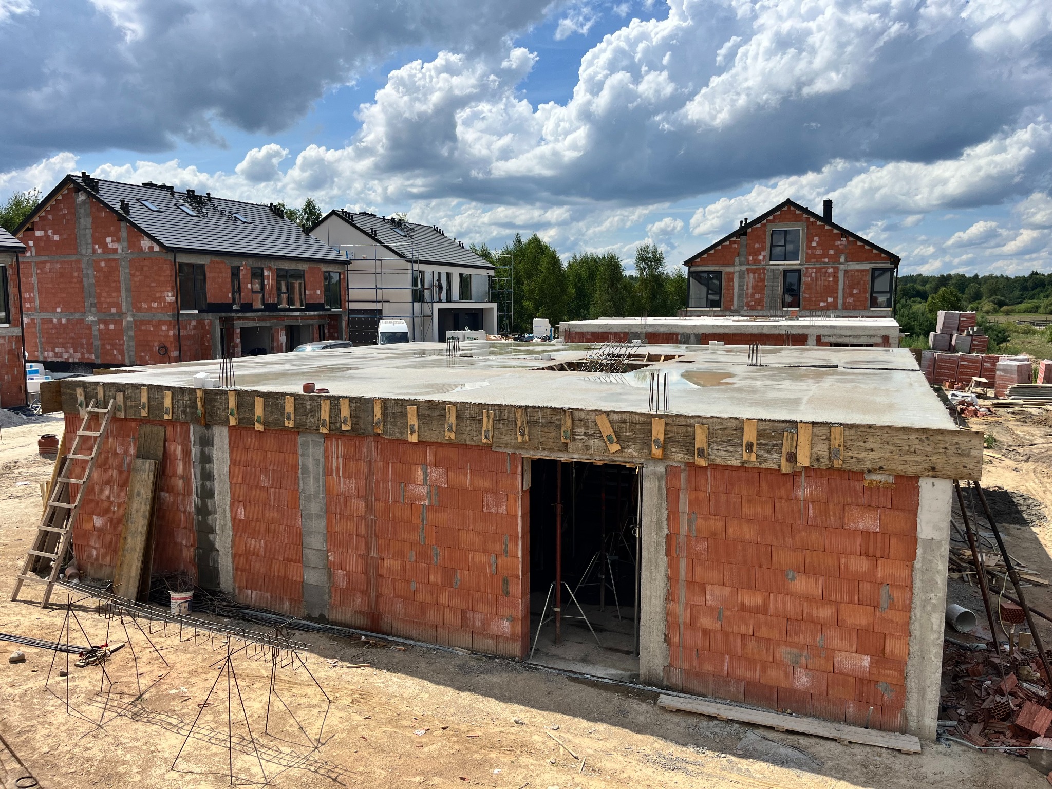 Surowy stan budowy domów jednorodzinnych z czerwonej cegły, widoczne zbrojenia na stropie, drewniane szalunki i drabina oparta o ścianę, w tle pochmurne niebo.