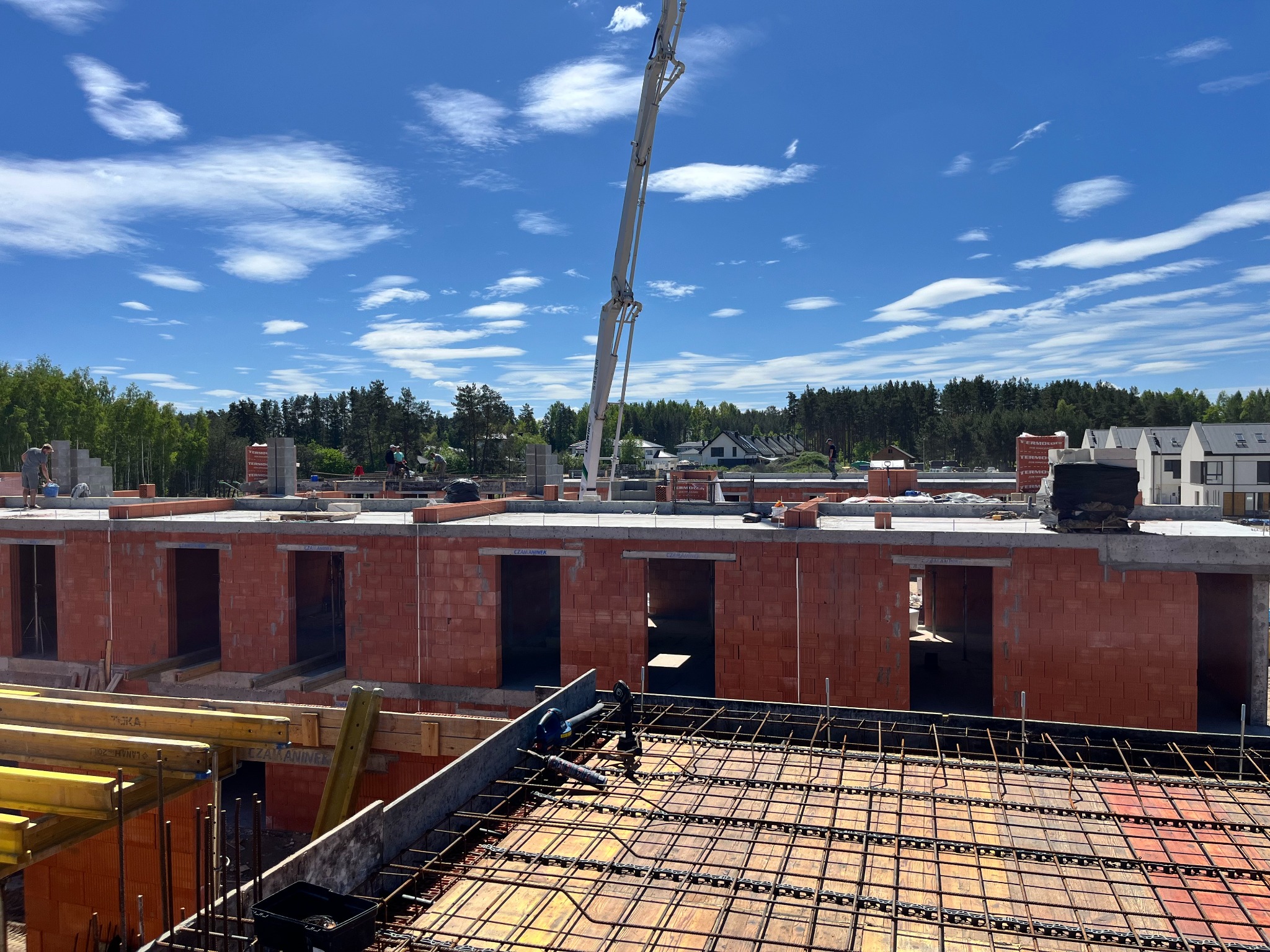 Budowa dwukondygnacyjnego budynku z czerwonej cegły ceramicznej, widoczna konstrukcja zbrojeniowa na pierwszym planie oraz pompa do betonu w tle, błękitne niebo z chmurami.