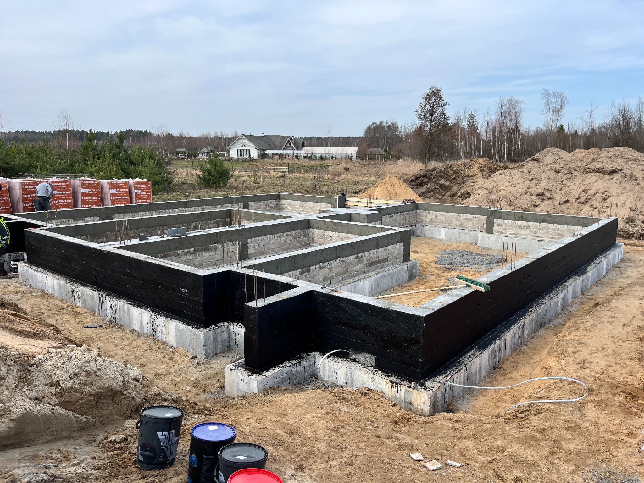 Fundamenty domu jednorodzinnego z izolacją przeciwwilgociową, widoczne puszki z preparatem izolacyjnym, w tle sterta cegieł i budynek mieszkalny.