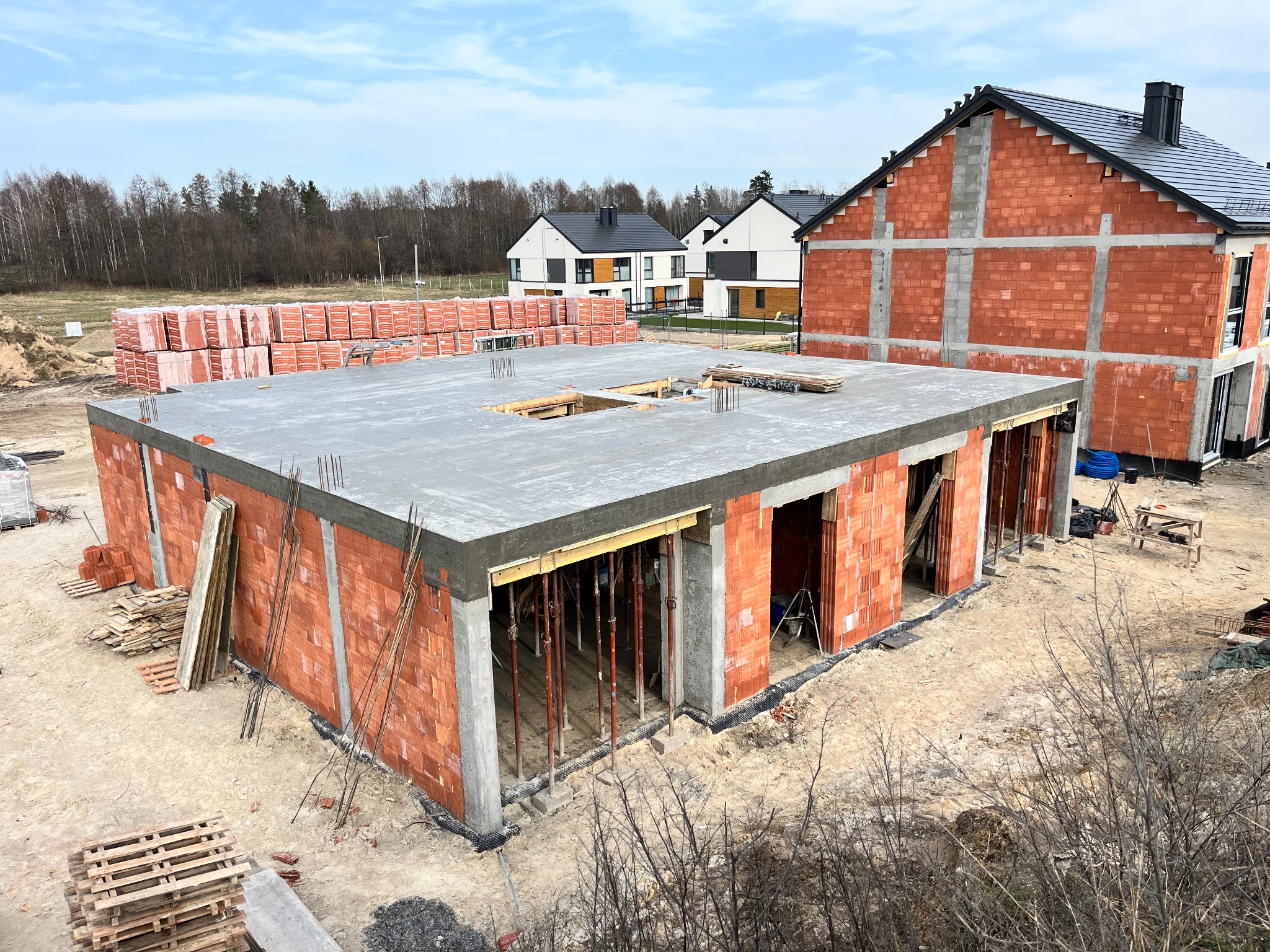 Stan surowy otwarty domu jednorodzinnego z czerwonej cegły, widoczne szalunki i betonowy strop, w tle inne domy w zabudowie szeregowej.