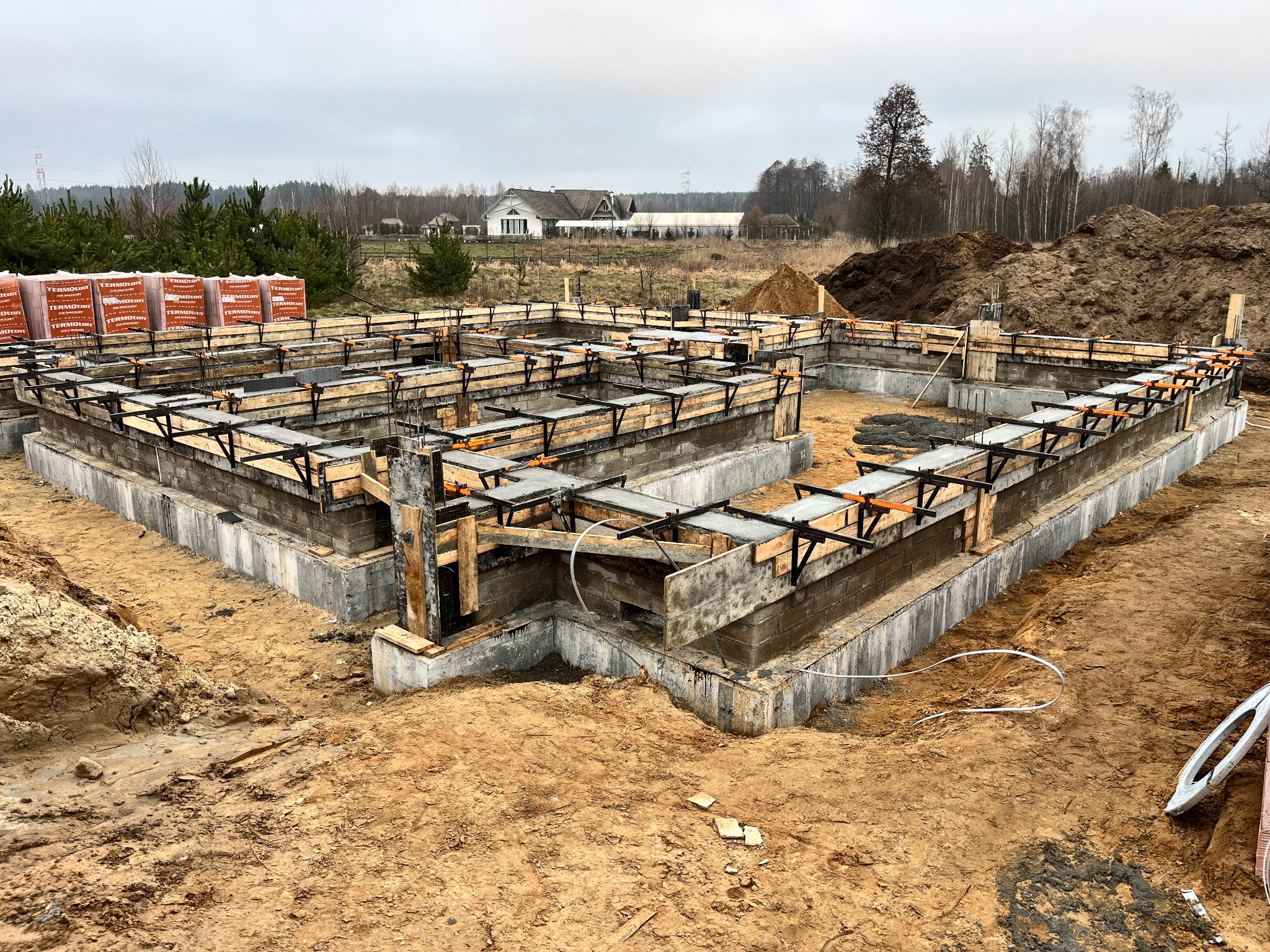 Wylany fundament z szalunkiem drewnianym i zbrojeniami, widoczne cegły na paletach, w tle dom jednorodzinny i drzewa.
