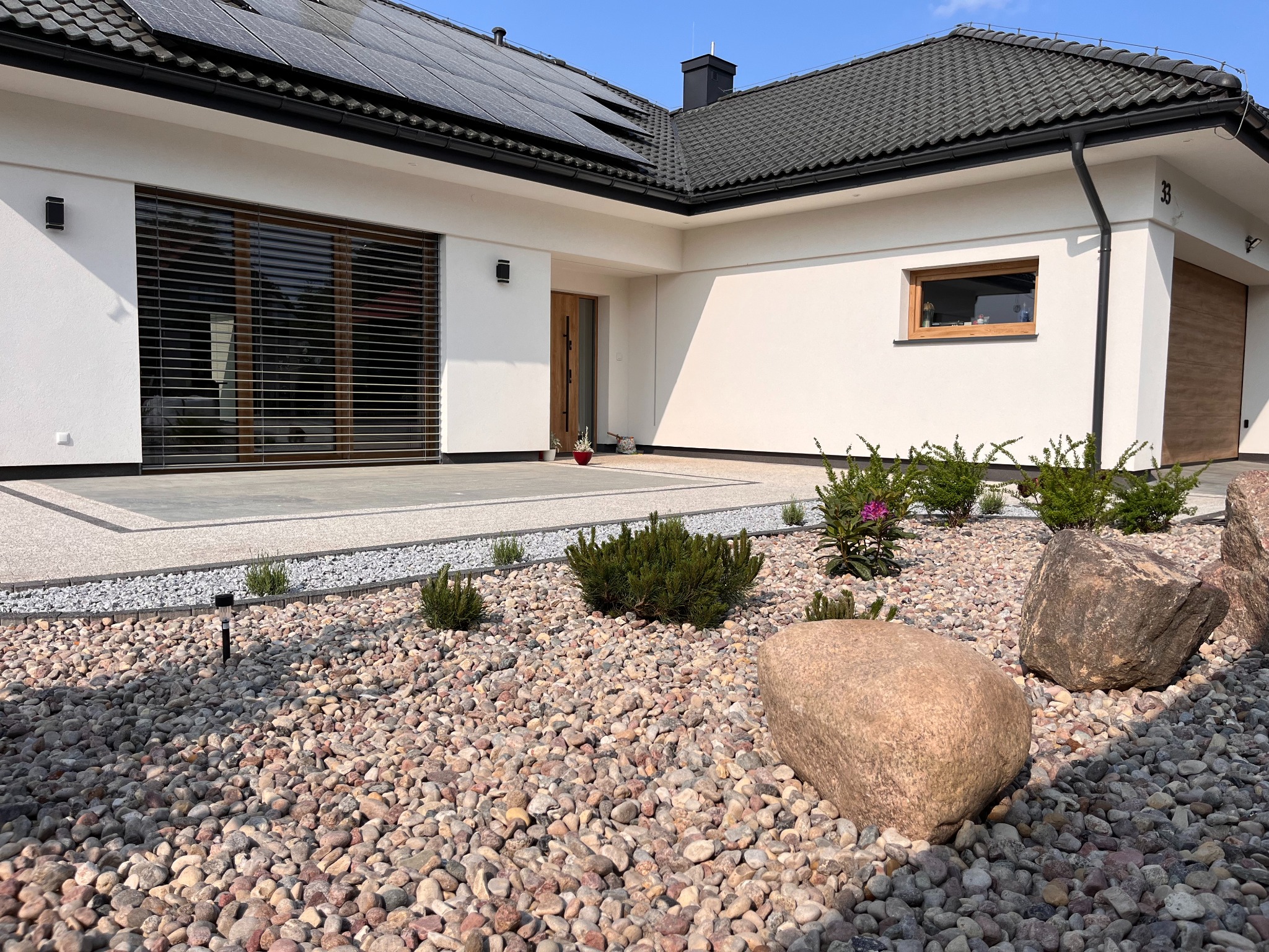 Nowoczesny dom jednorodzinny z panelami słonecznymi na dachu, jasną elewacją, drewnianymi oknami i żaluzjami zewnętrznymi, otoczony kamiennym ogrodem z krzewami i dużymi głazami.