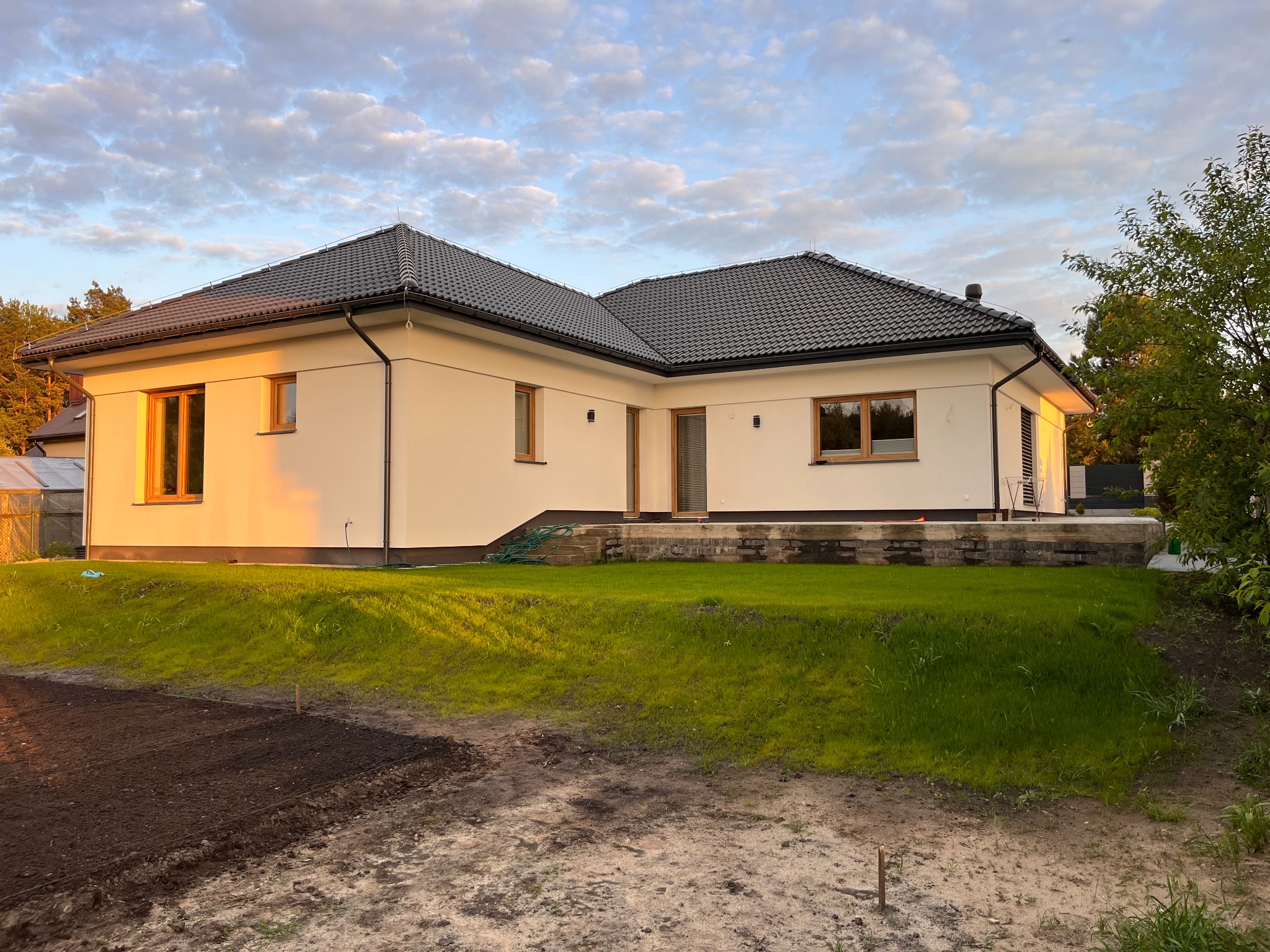 Nowoczesny, parterowy dom jednorodzinny z ciemną dachówką, oświetlony popołudniowym słońcem, z widocznym przygotowanym terenem pod ogródek.