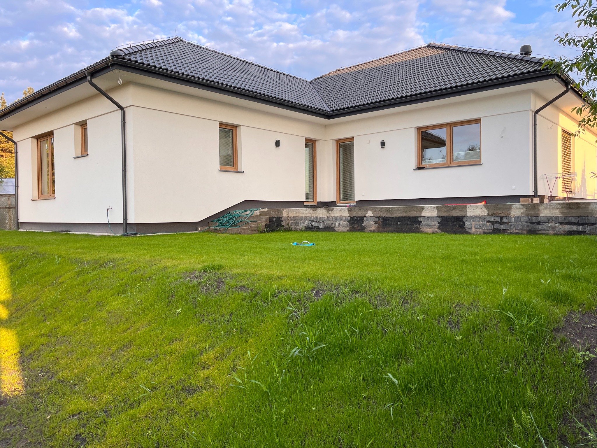 Nowoczesny dom jednorodzinny z białymi ścianami, ciemną dachówką i drewnianymi oknami, widok z trawnika z elementami prac wykończeniowych na tarasie.