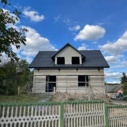 Dom w trakcie budowy, widoczne rusztowania, ściany z pustaków, czarny dach z dachówki, błękitne niebo z białymi chmurami, ogrodzenie z betonu i zielonej farby.