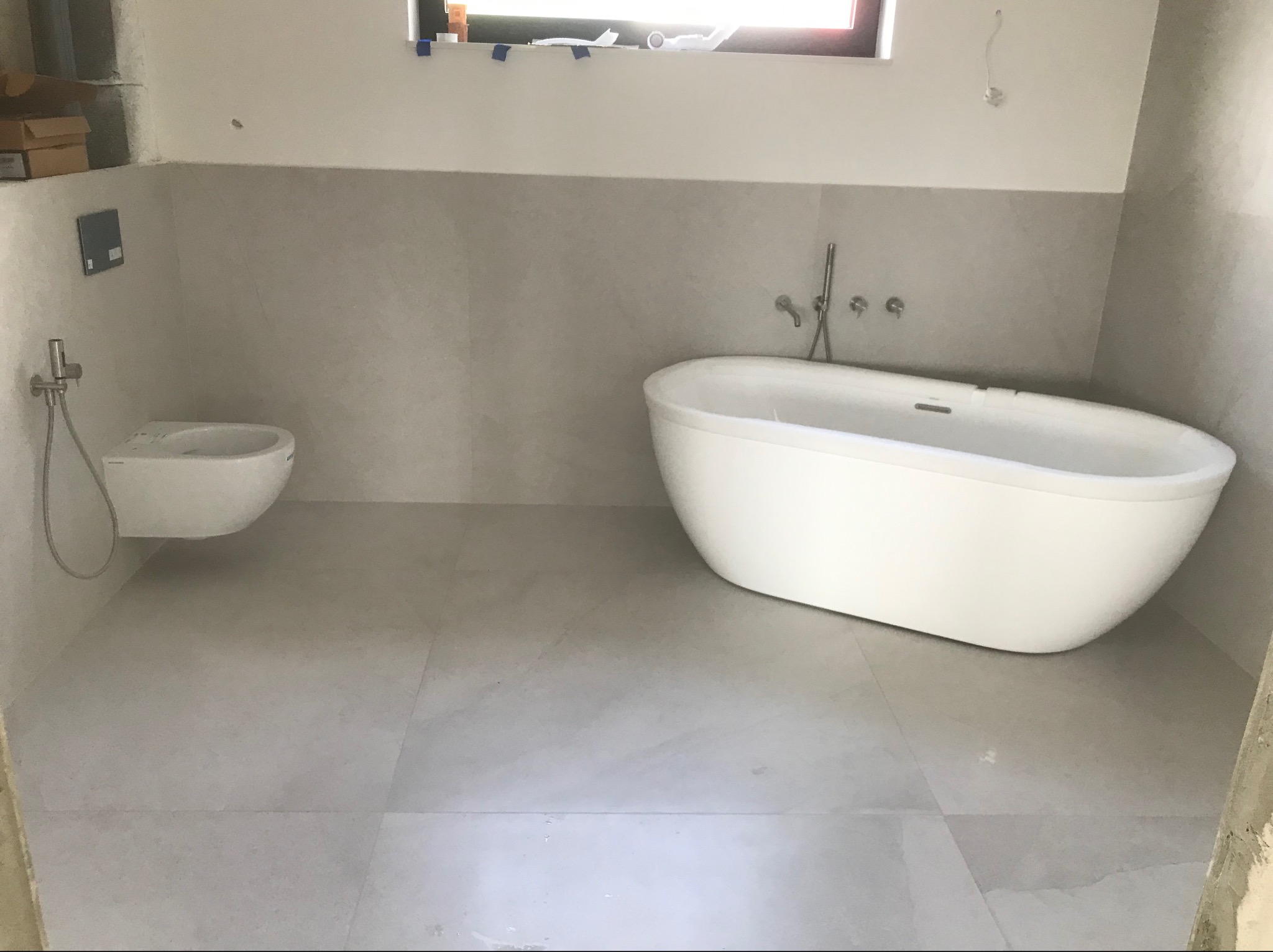 Nowoczesna, minimalistyczna łazienka w odcieniach szarości z wolnostojącą białą wanną i podwieszaną toaletą z chromowaną armaturą, okno z niebieską taśmą malarską u góry.