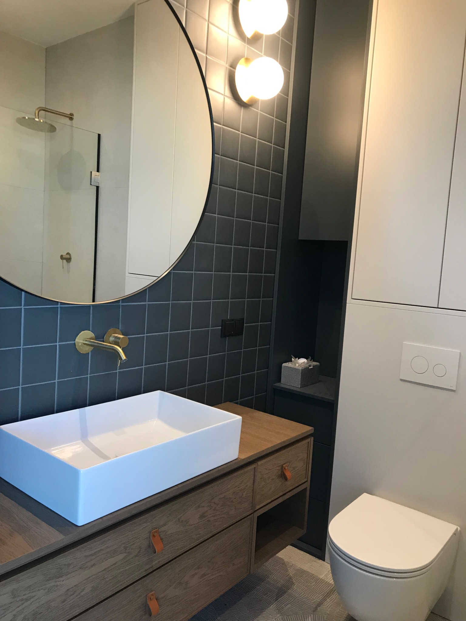 Nowoczesna łazienka z umywalką nablatową na drewnianej szafce z skórzanymi uchwytami, okrągłe lustro z podświetleniem, szare płytki na ścianie, minimalistyczna toaleta podtynkowa i kabina...