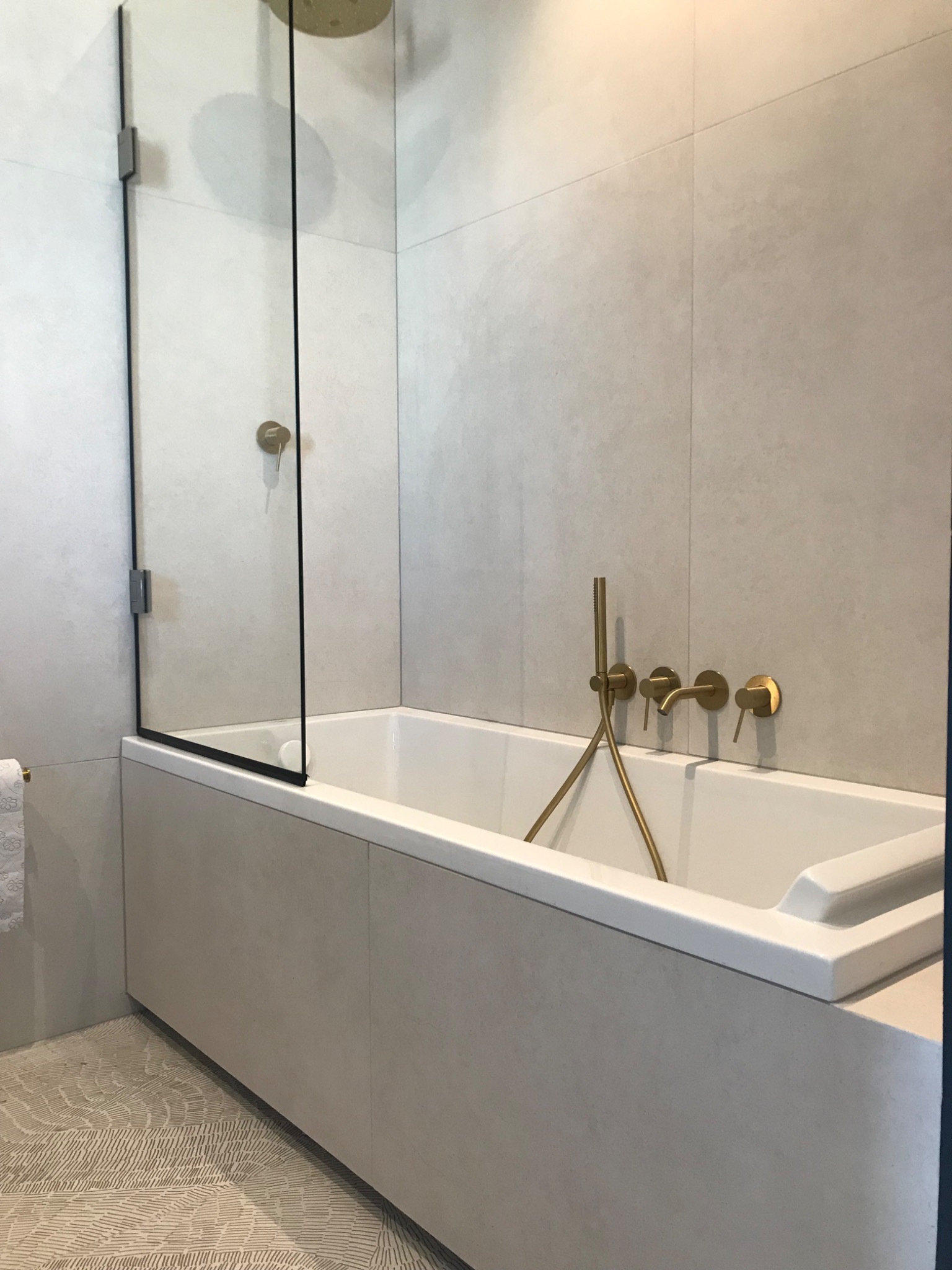 Nowoczesna łazienka z prostokątną wanną obudowaną płytami w kolorze beżowym, z zamontowaną szklaną ścianką prysznicową i złotą armaturą.