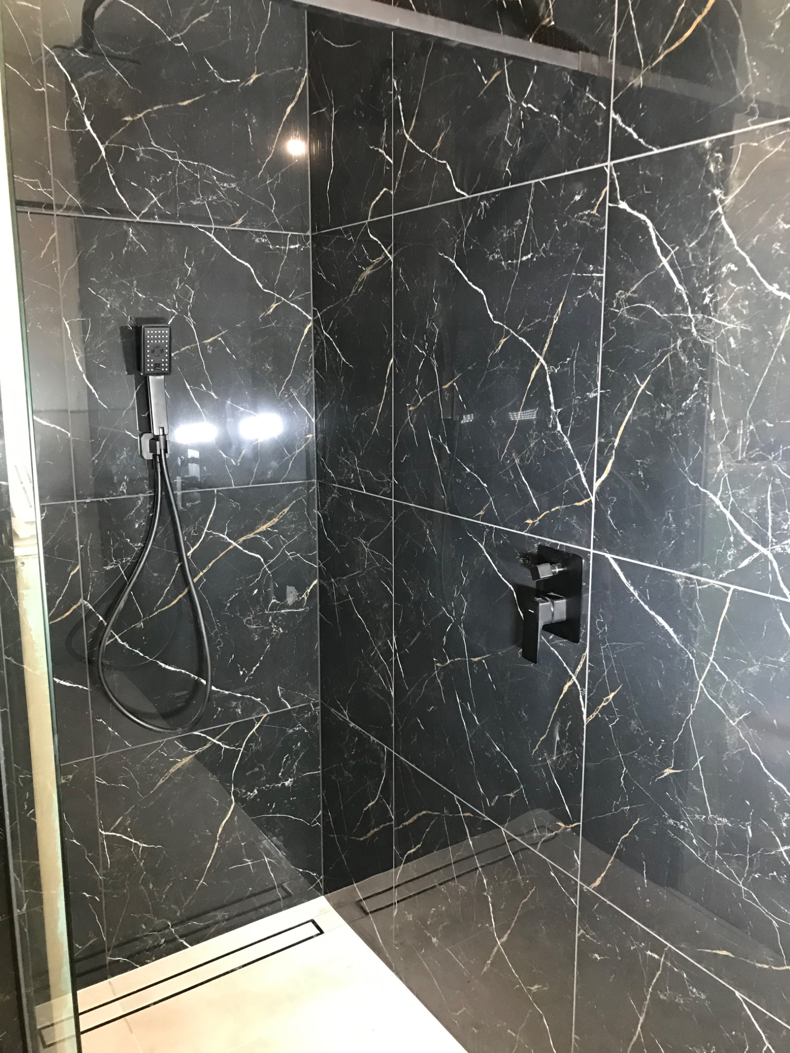 Nowoczesna kabina prysznicowa z czarnymi płytkami imitującymi marmur z białymi i złotymi żyłami, wyposażona w czarną armaturę i odpływ liniowy.