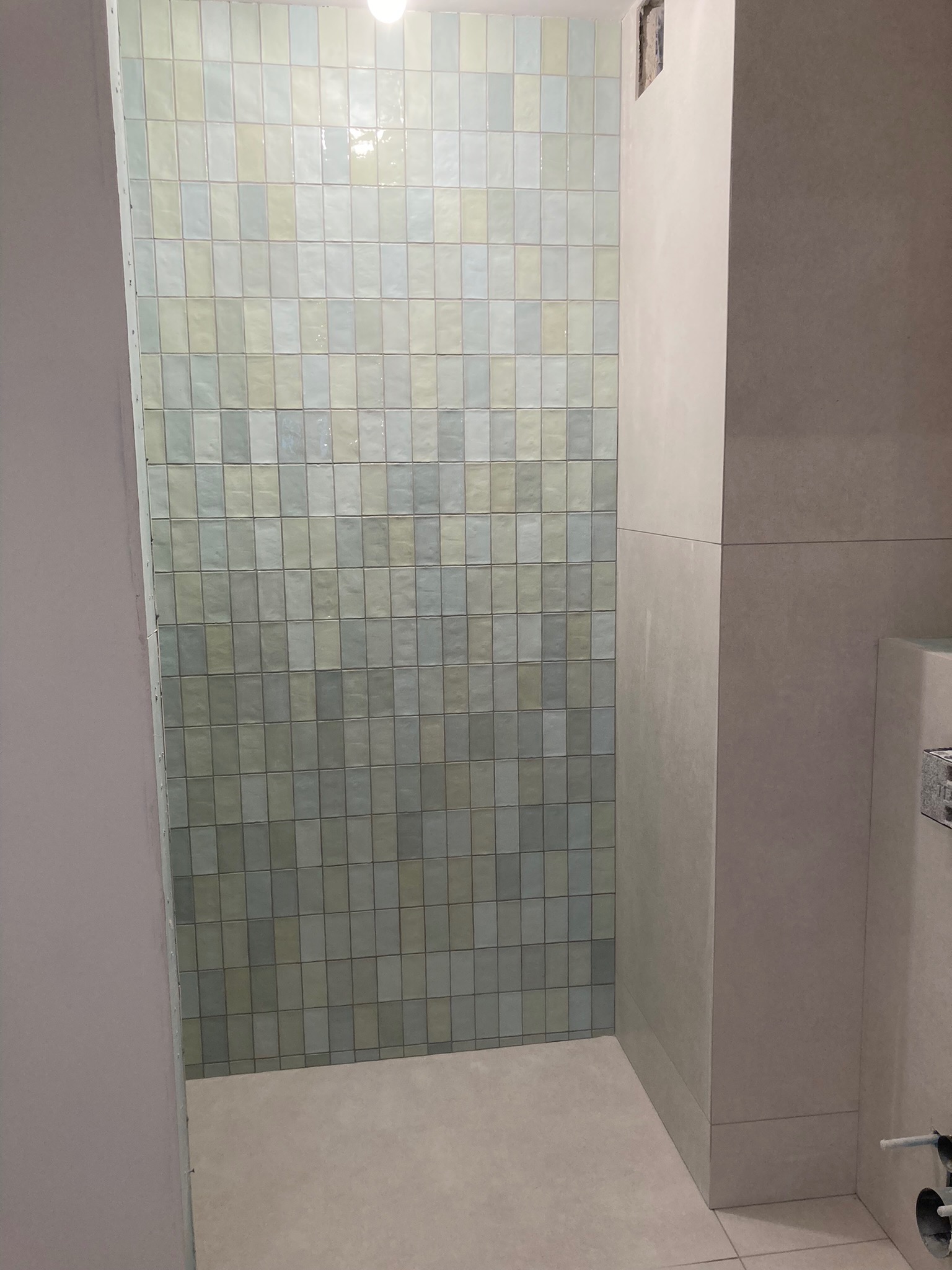 Wykończona ściana w łazience z pionowo ułożonymi, błyszczącymi płytkami w odcieniach szarości, zieleni i bieli, sąsiadująca z gładką, pomalowaną ścianą. Widoczne fragmenty instalacji wodnej.