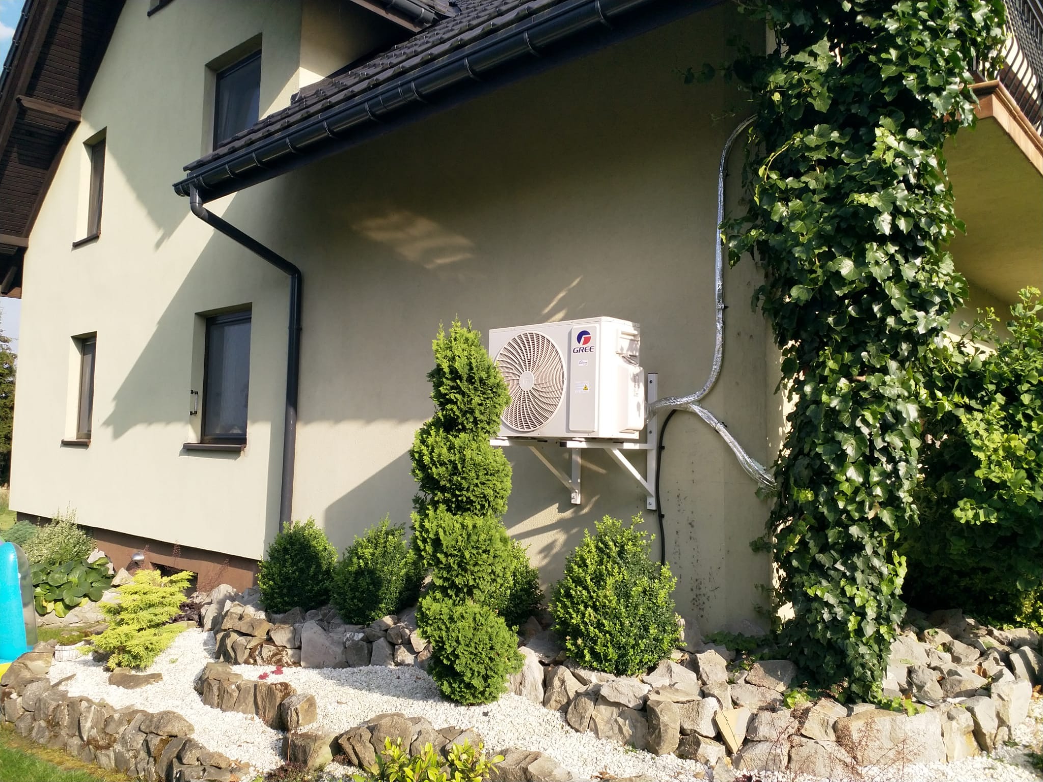 Zewnętrzna jednostka klimatyzatora GREE zamontowana na elewacji domu, z widocznymi przewodami w izolacji, na tle dekoracyjnego skalniaka z białym grysem i ozdobnymi krzewami.