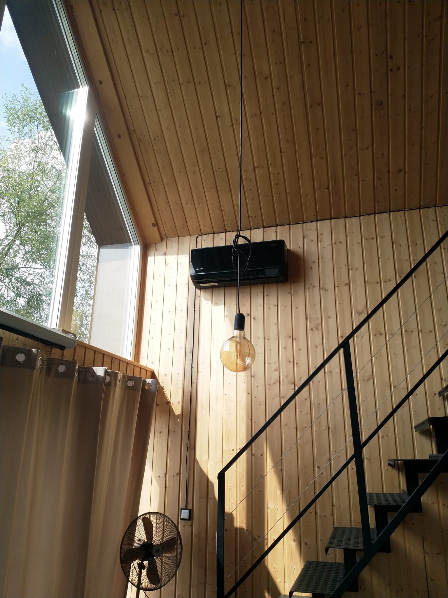 Wnętrze domu z drewnianą boazerią, klimatyzatorem ściennym, czarnymi schodami i wiszącą żarówką w stylu retro, oświetlone naturalnym światłem wpadającym przez okno.