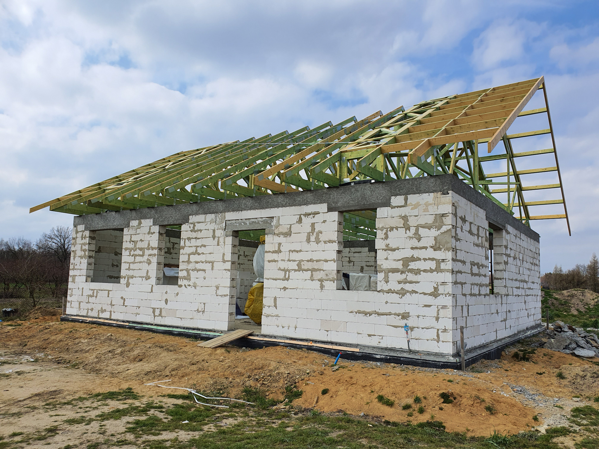 Surowy stan budynku z białych bloczków z częściowo zamontowaną drewnianą konstrukcją dachu pod błękitnym niebem.