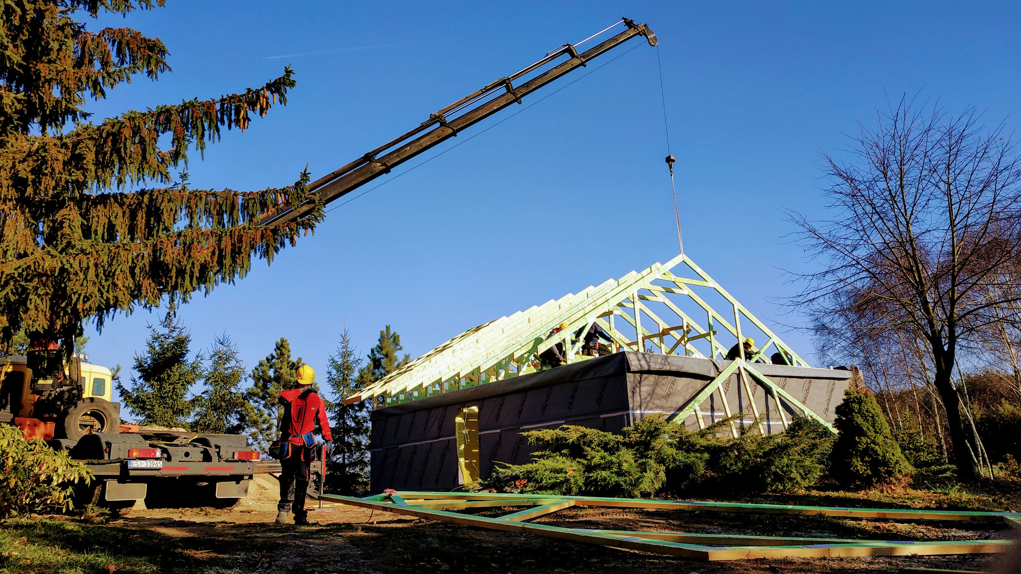 Montaż drewnianej konstrukcji dachu domu jednorodzinnego z użyciem dźwigu, w tle las iglasty i pracownik w czerwonej kurtce nadzorujący prace.