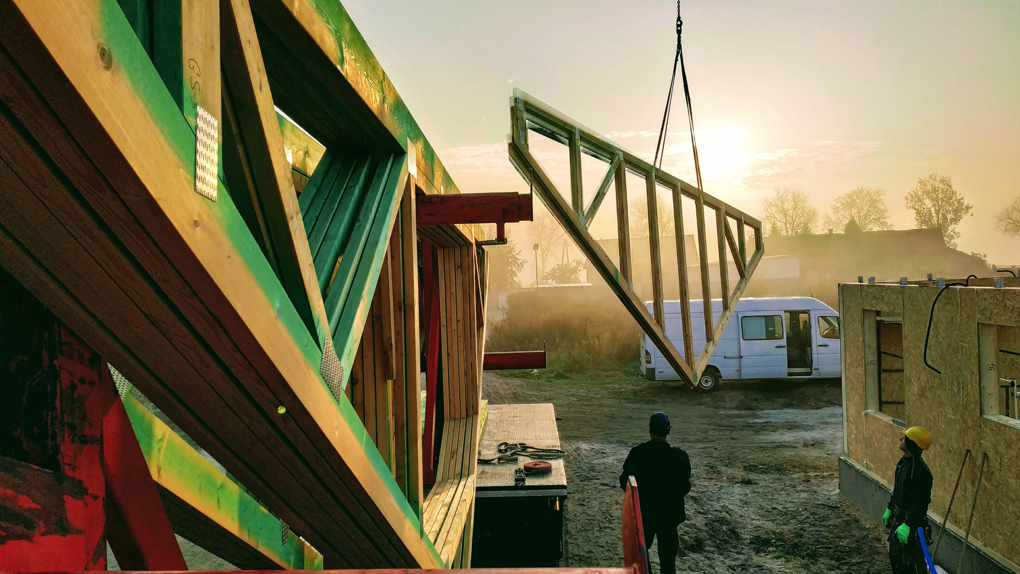 Montaż drewnianej konstrukcji szkieletowej domu jednorodzinnego; widoczny dźwig podnoszący element ramy na tle porannej mgły i zaparkowanego białego vana.