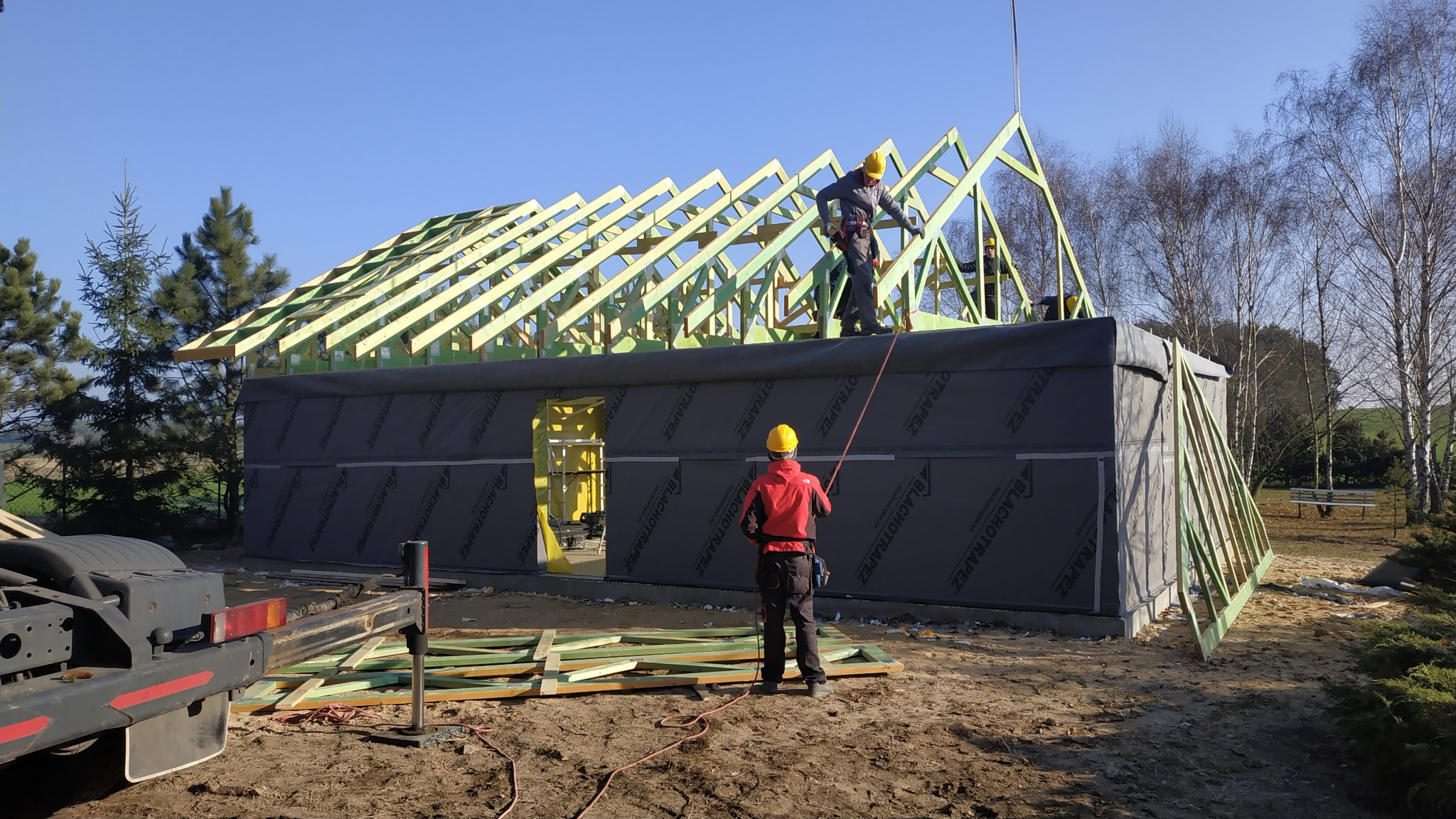 Montaż drewnianej konstrukcji dachu na budynku z czarną membraną, dwóch pracowników w kaskach, jeden na dachu, drugi na ziemi, widoczny żuraw samochodowy w tle.