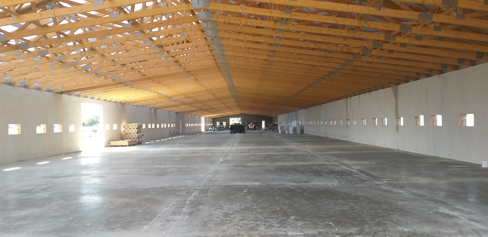 Wnętrze hali w trakcie budowy z drewnianą konstrukcją dachu, betonową posadzką i rzędami małych okien w ścianach, widoczne materiały izolacyjne i ciężki sprzęt w tle.