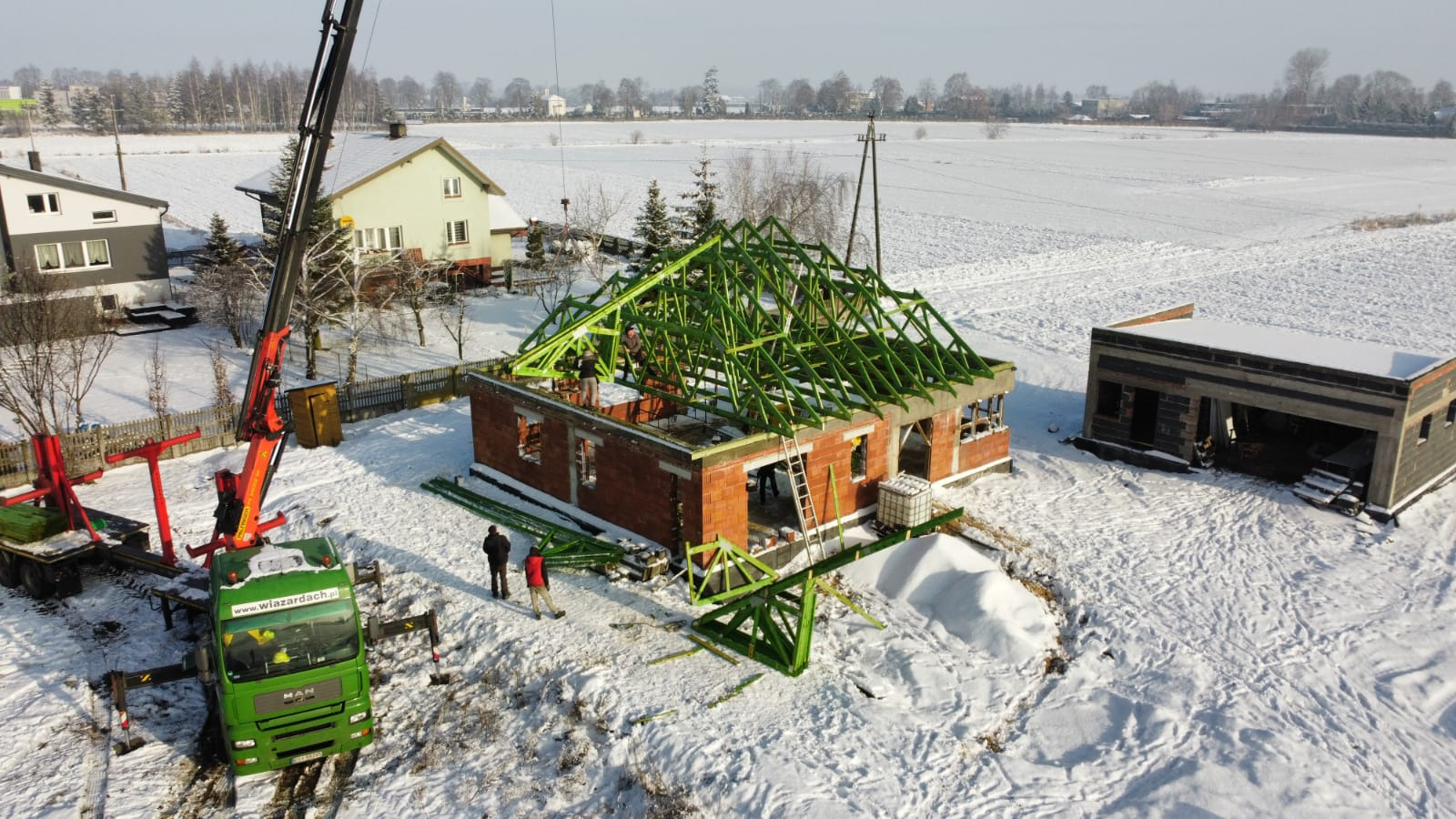 Montaż zielonej więźby dachowej na surowym budynku z czerwonej cegły w zimowej scenerii, z użyciem dźwigu i obecnością pracowników na dachu oraz na śniegu.