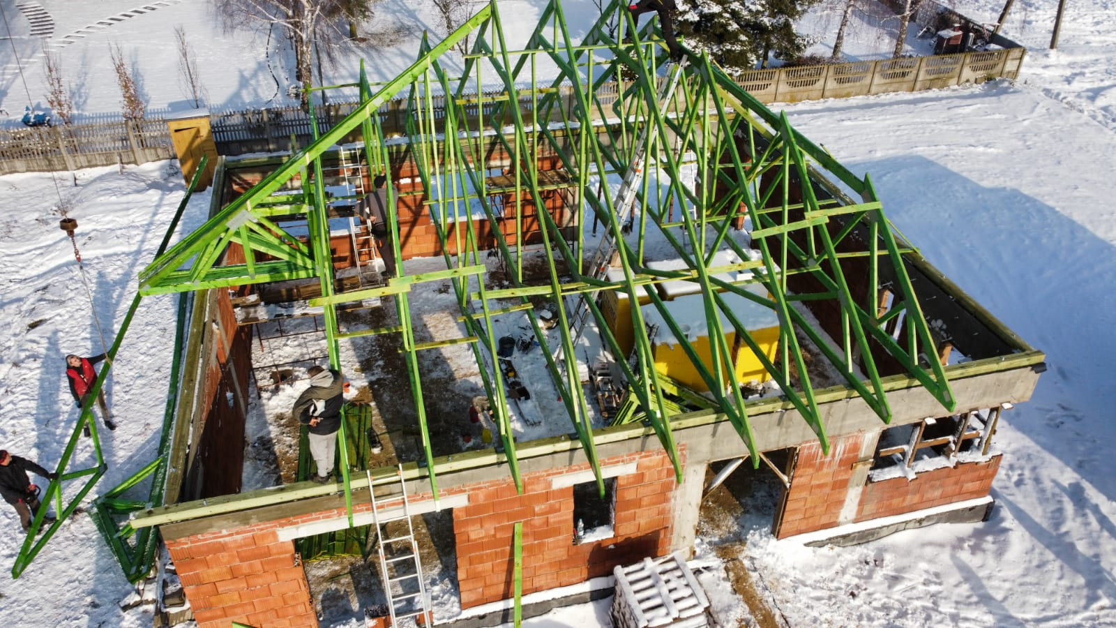 Budowa domu jednorodzinnego w zimowej scenerii: widok z góry na murowane ściany z czerwonej cegły i zieloną konstrukcję drewnianą dachu, z pracownikami w trakcie montażu.