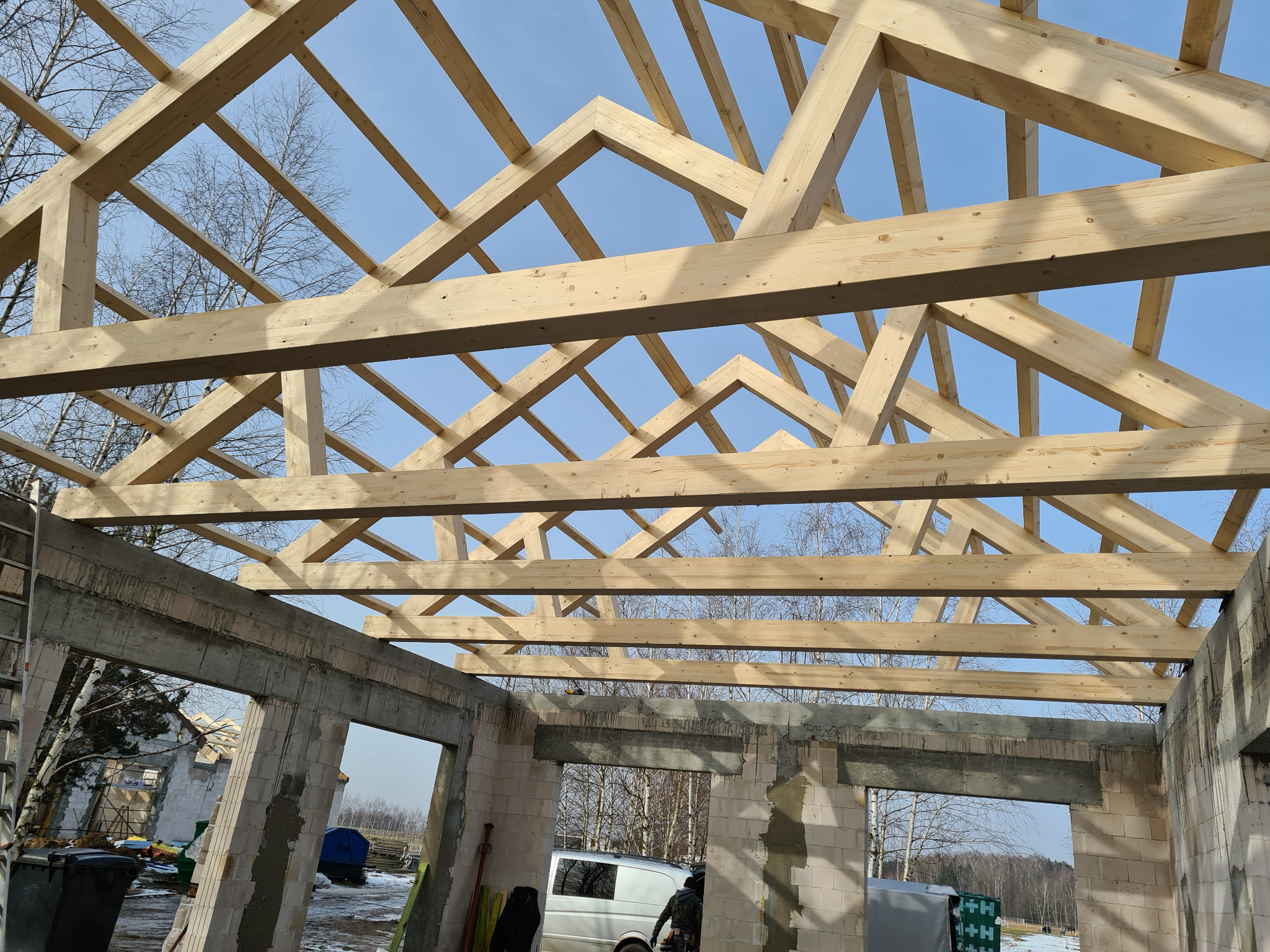Konstrukcja drewniana dachu w trakcie budowy, widok z wnętrza budynku z betonu komórkowego, na tle błękitnego nieba i drzew bez liści.
