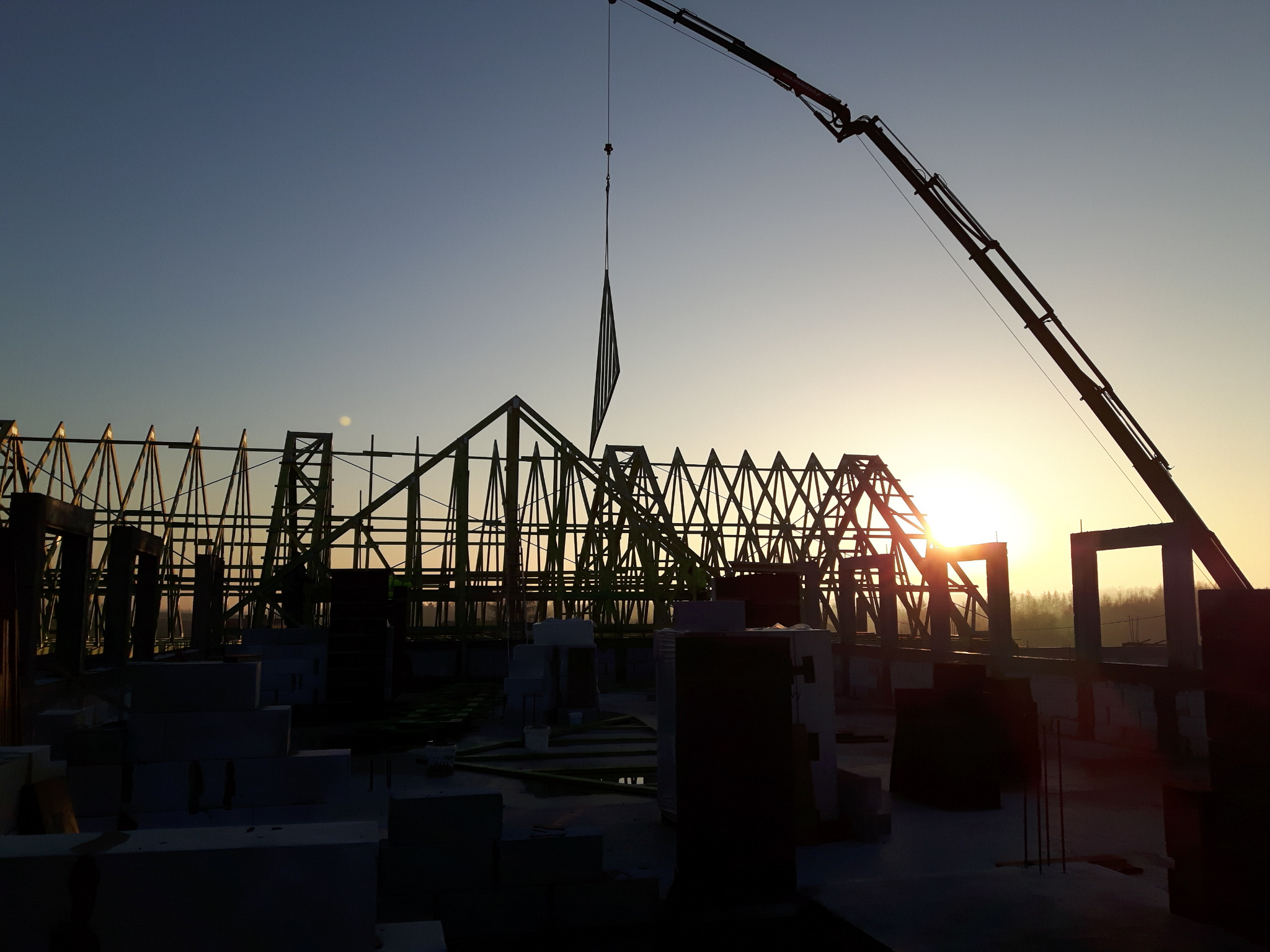 Konstrukcja drewniana dachu w trakcie montażu, podnoszona przez dźwig na tle wschodzącego słońca, widoczne bloczki betonowe.