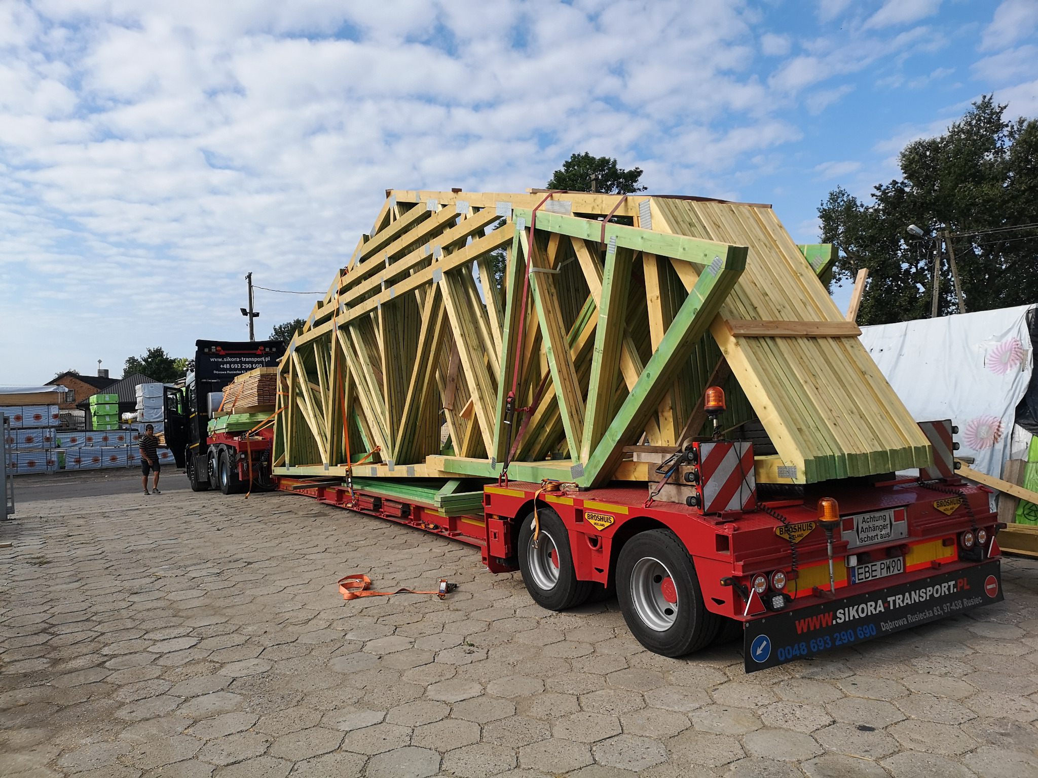 Transport konstrukcji drewnianej dachu na czerwonej platformie transportowej firmy Sikora Transport, widok z tyłu, w tle składowane materiały budowlane i pracownik.