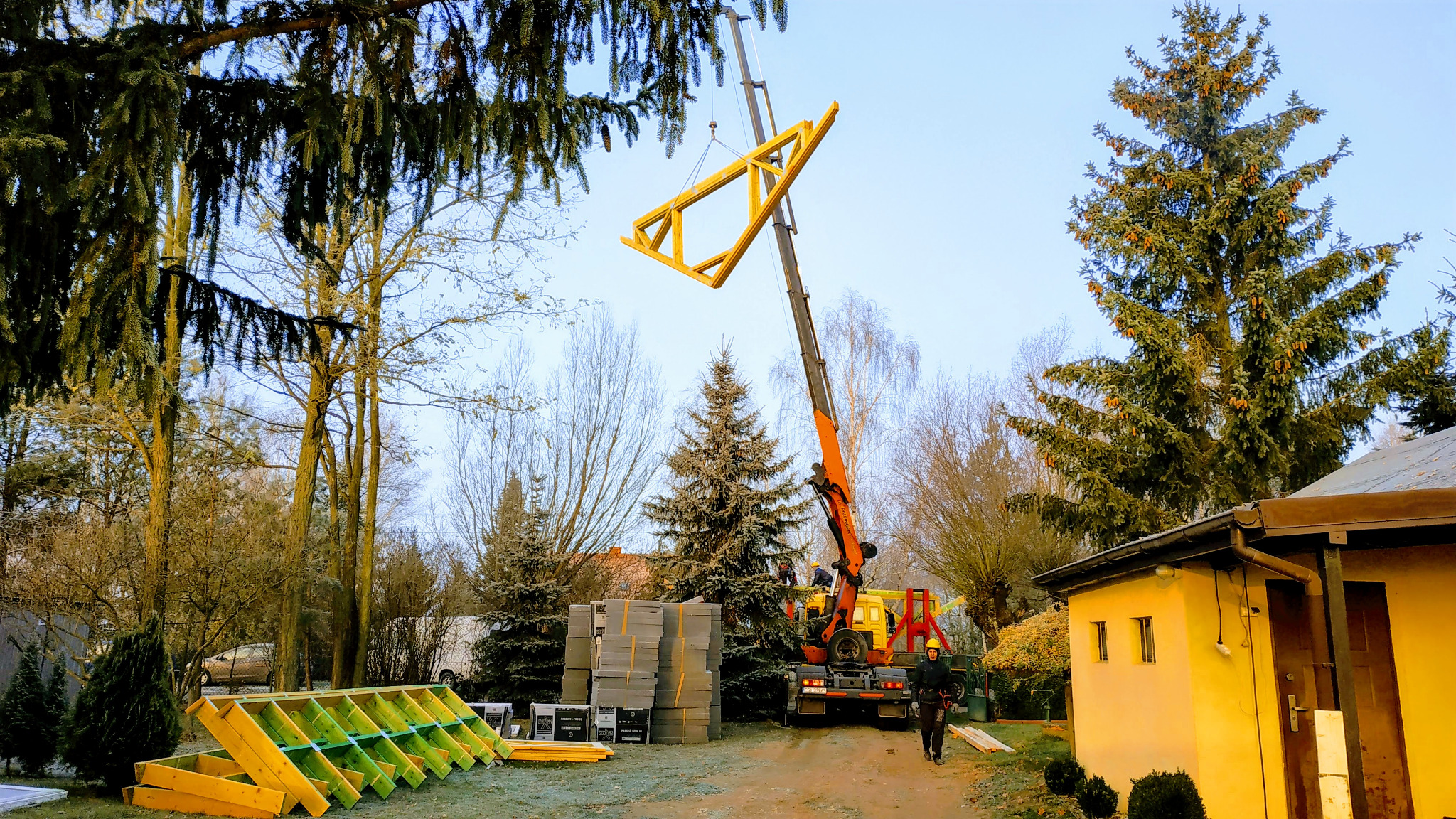 Żółta drewniana konstrukcja dachu podnoszona przez pomarańczowy dźwig na placu budowy z widocznymi elementami konstrukcyjnymi, bloczkami betonowymi i pracownikiem w oddali.