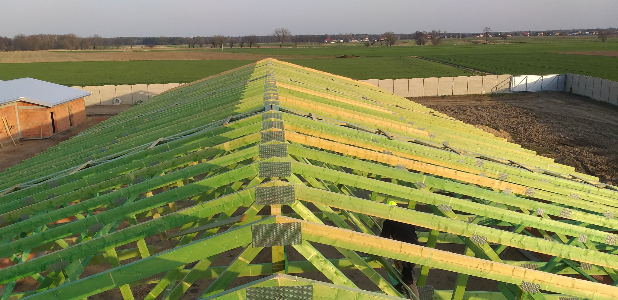 Wysoki kąt widzenia na konstrukcję dachu z impregnowanego drewna w kolorze zielonym i żółtym, z widocznymi łącznikami metalowymi, w tle krajobraz polny i budynek w trakcie budowy.