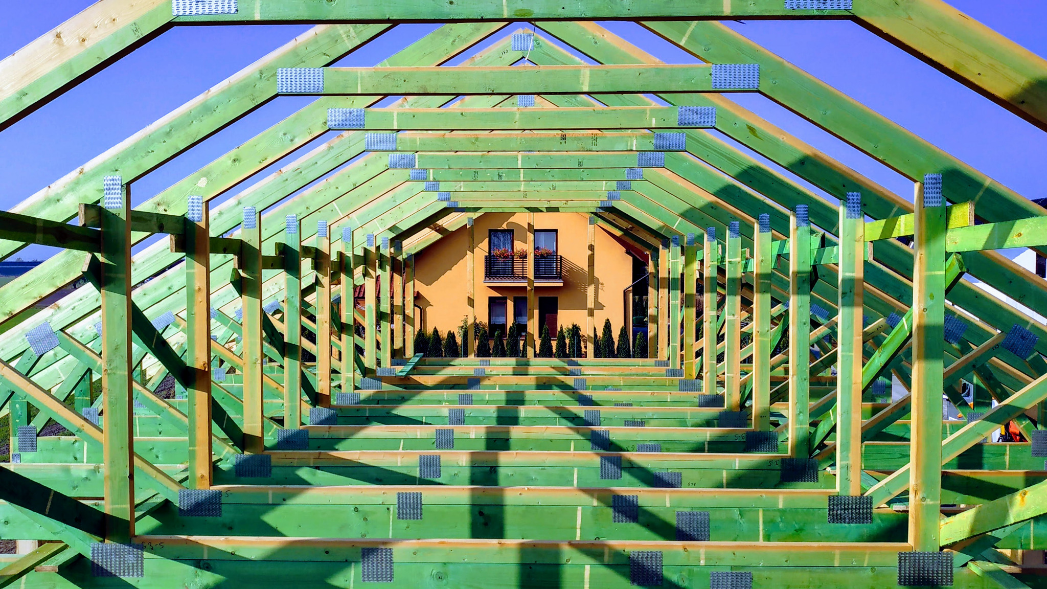 Zielona konstrukcja drewniana dachu z metalowymi łącznikami, w tle widoczny pomarańczowy dom z balkonem i tujami.