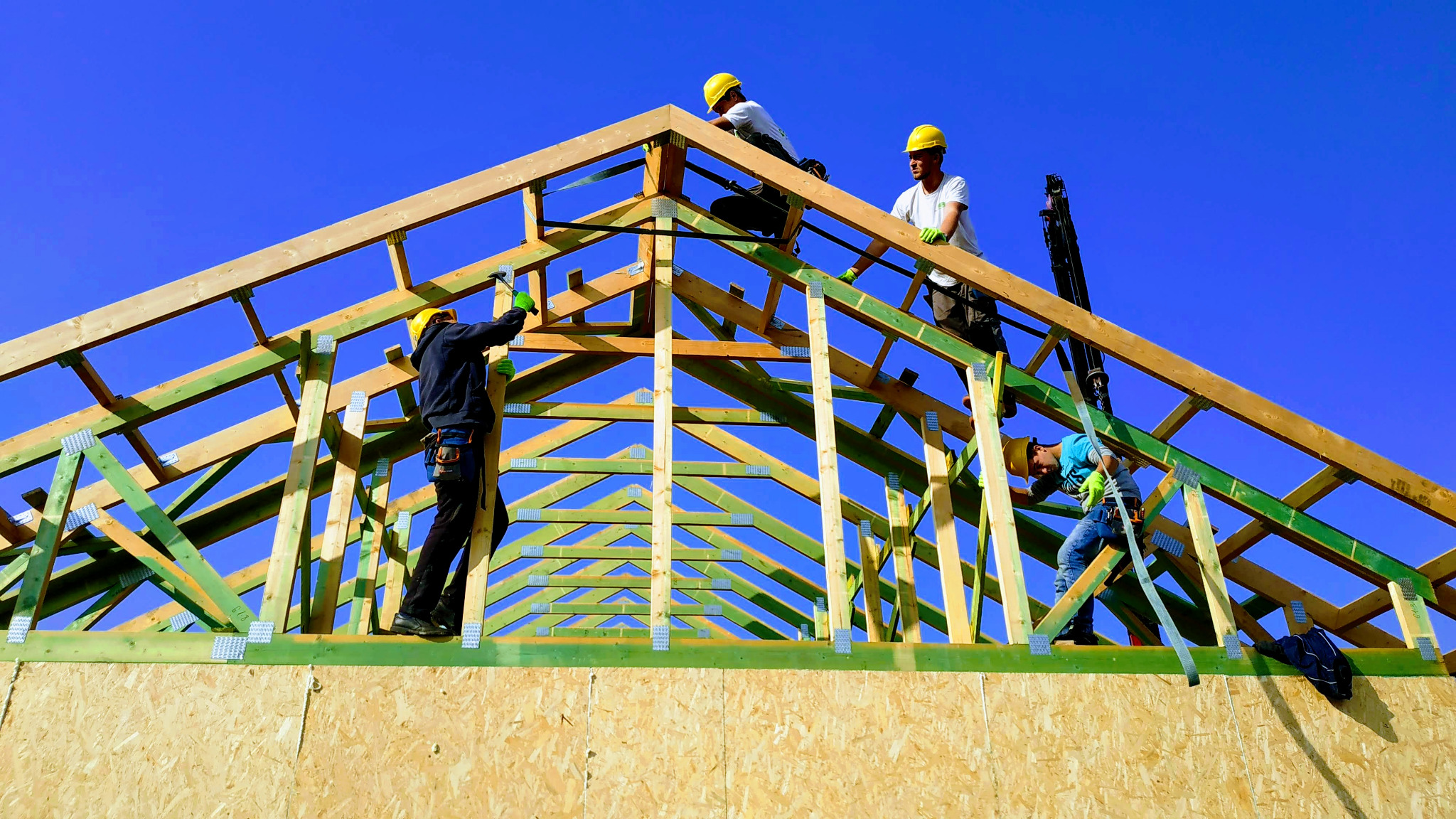 Czterech robotników w żółtych kaskach montuje drewnianą konstrukcję dachu na tle błękitnego nieba. Widoczne elementy konstrukcyjne pomalowane na zielono.