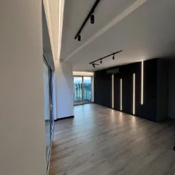 Nowoczesne wnętrze z podłogą z paneli, czarną ścianą z pionowymi pasami światła LED i szynoprzewodem z reflektorami punktowymi na suficie.